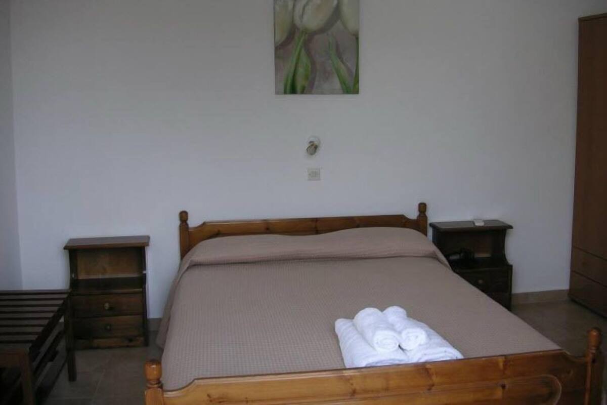 A single bed with a wooden frame, a beige bedspread, and a set of rolled white towels on the footboard. There is a small wooden nightstand to the left and a larger one to the right of the bed. A painting of tulips hangs on the wall above the bed.