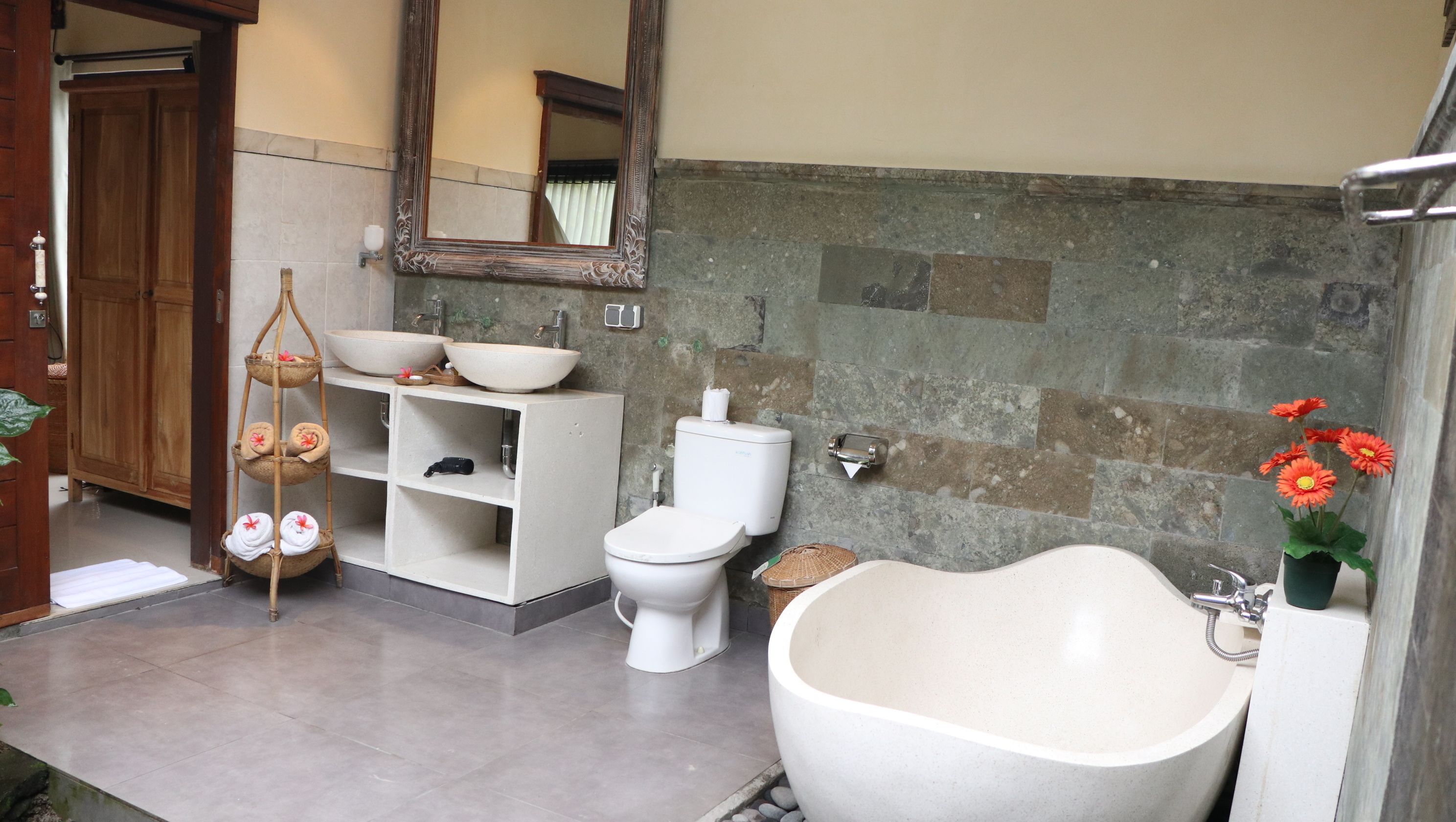 Un baño con una bañera independiente grande, dos lavamanos con espejo arriba, un inodoro y una pared de piedra.