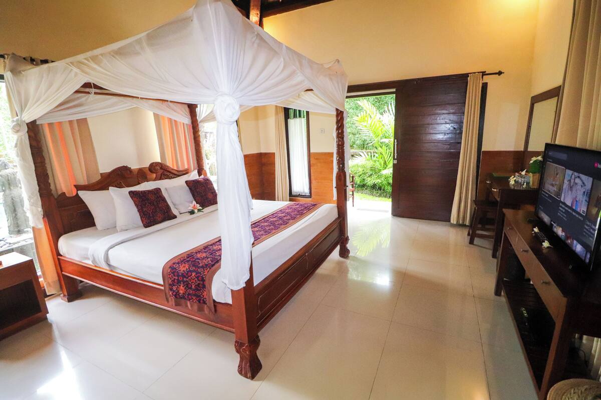 A four-poster bed with a canopy, a television on a wooden stand, and a view of the outdoors through a door.