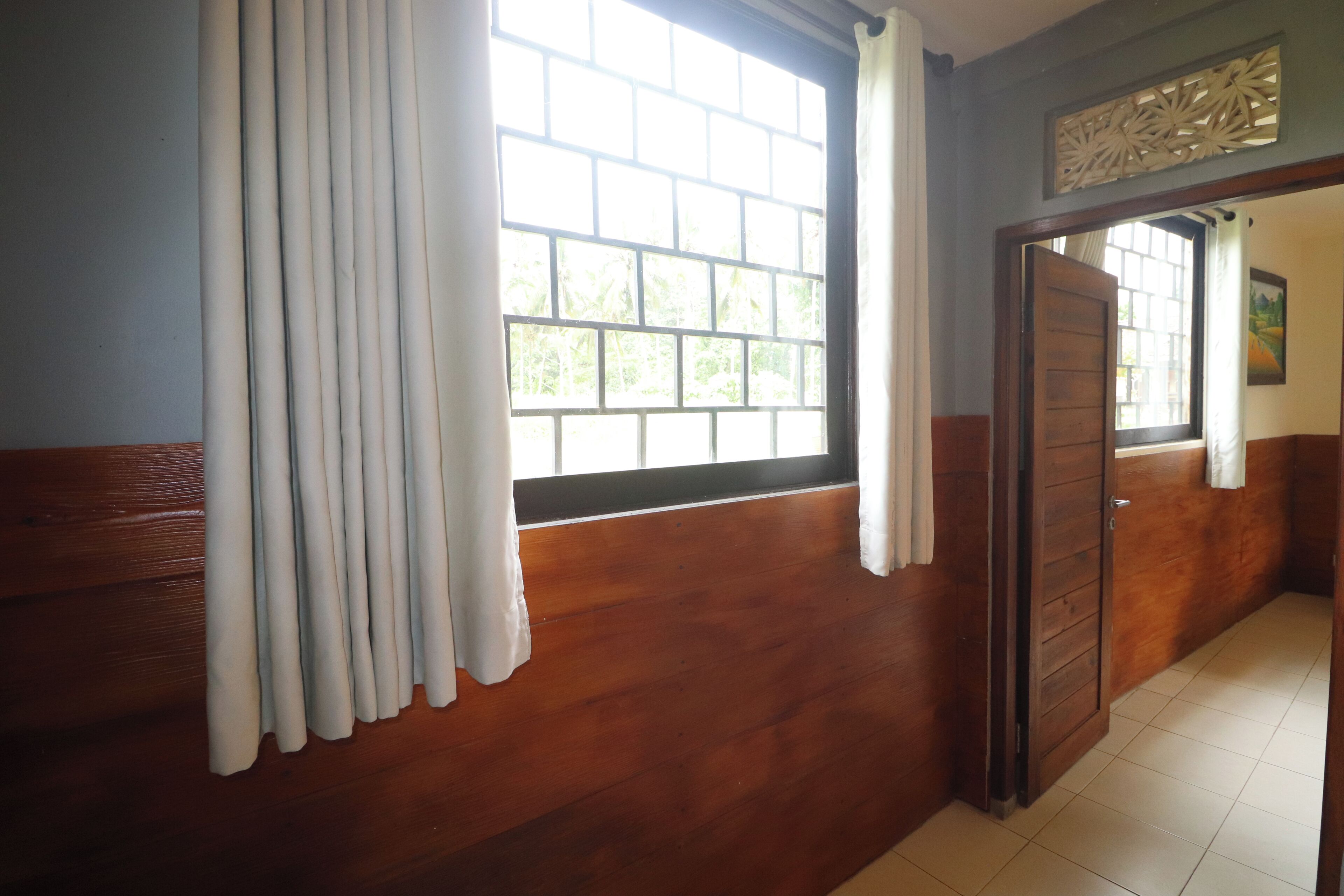 A room with wooden walls, a large window with white curtains, a door, and a tiled floor.
