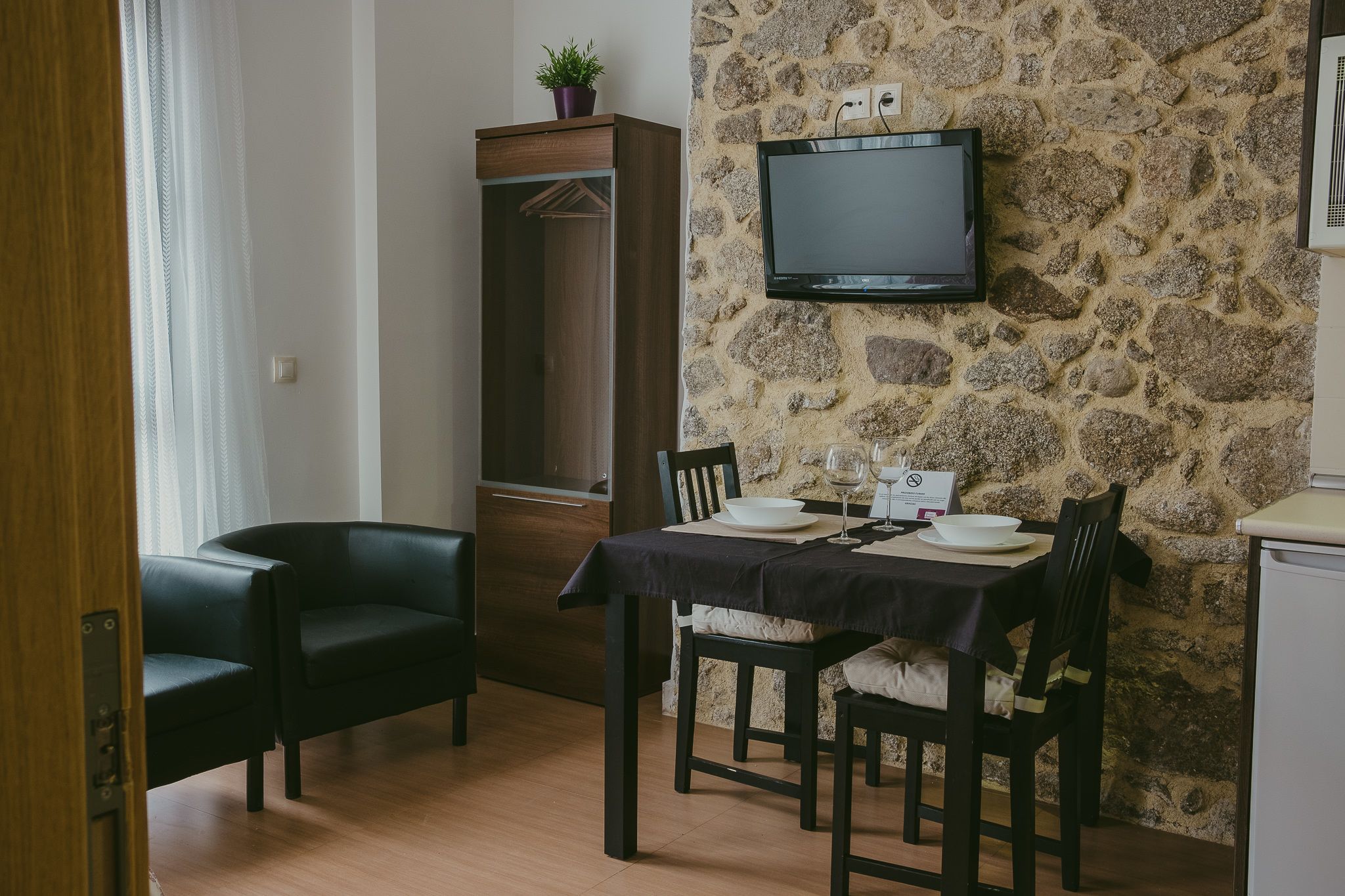 A dining area with a stone wall, a flat-screen TV mounted above a table set for two, and a wooden cabinet with a plant on top.