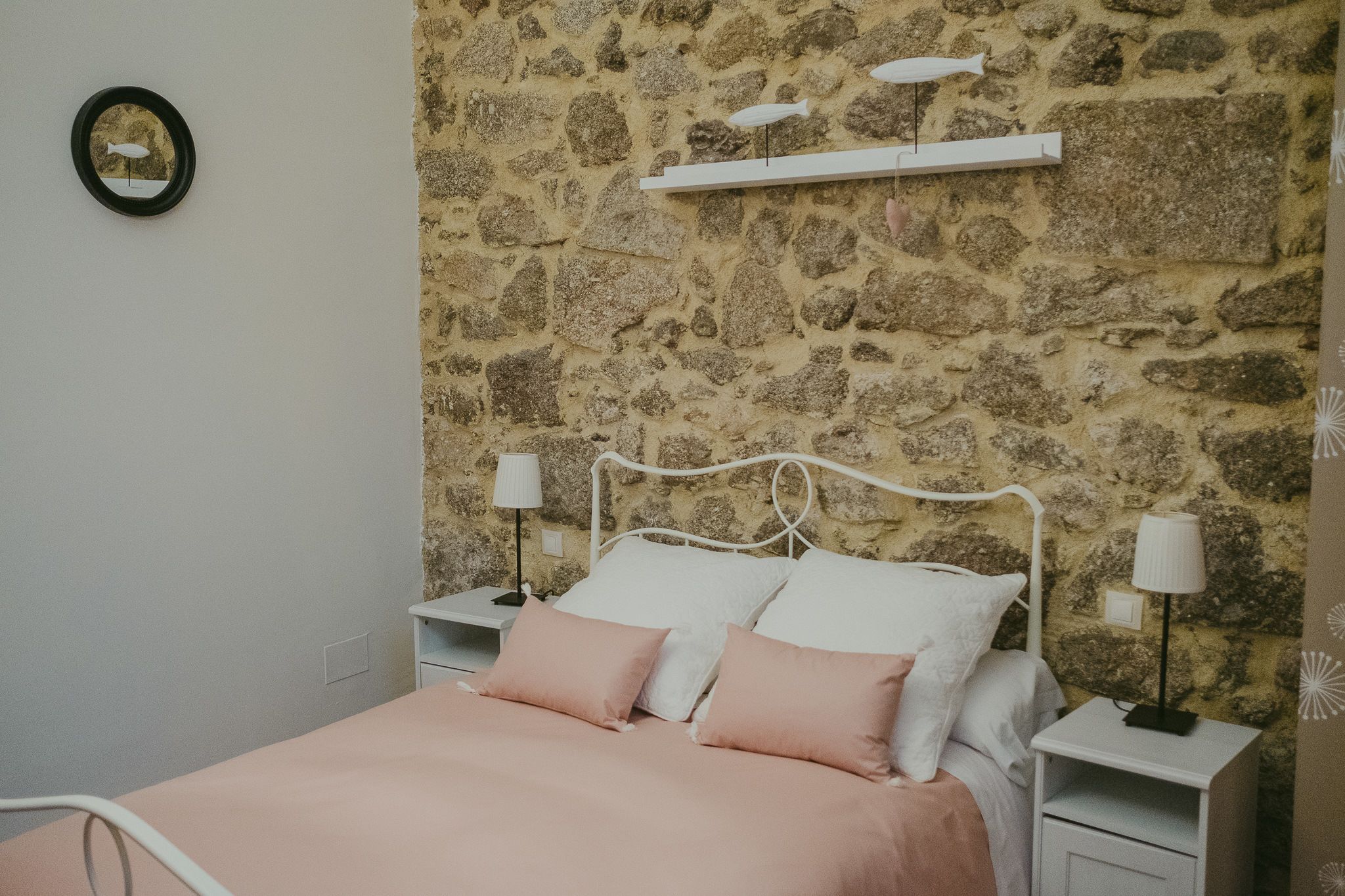 Un dormitorio con pared de piedra, cama con ropa de cama blanca y rosa, dos mesitas de noche con lámparas y un pequeño estante sobre la cama.