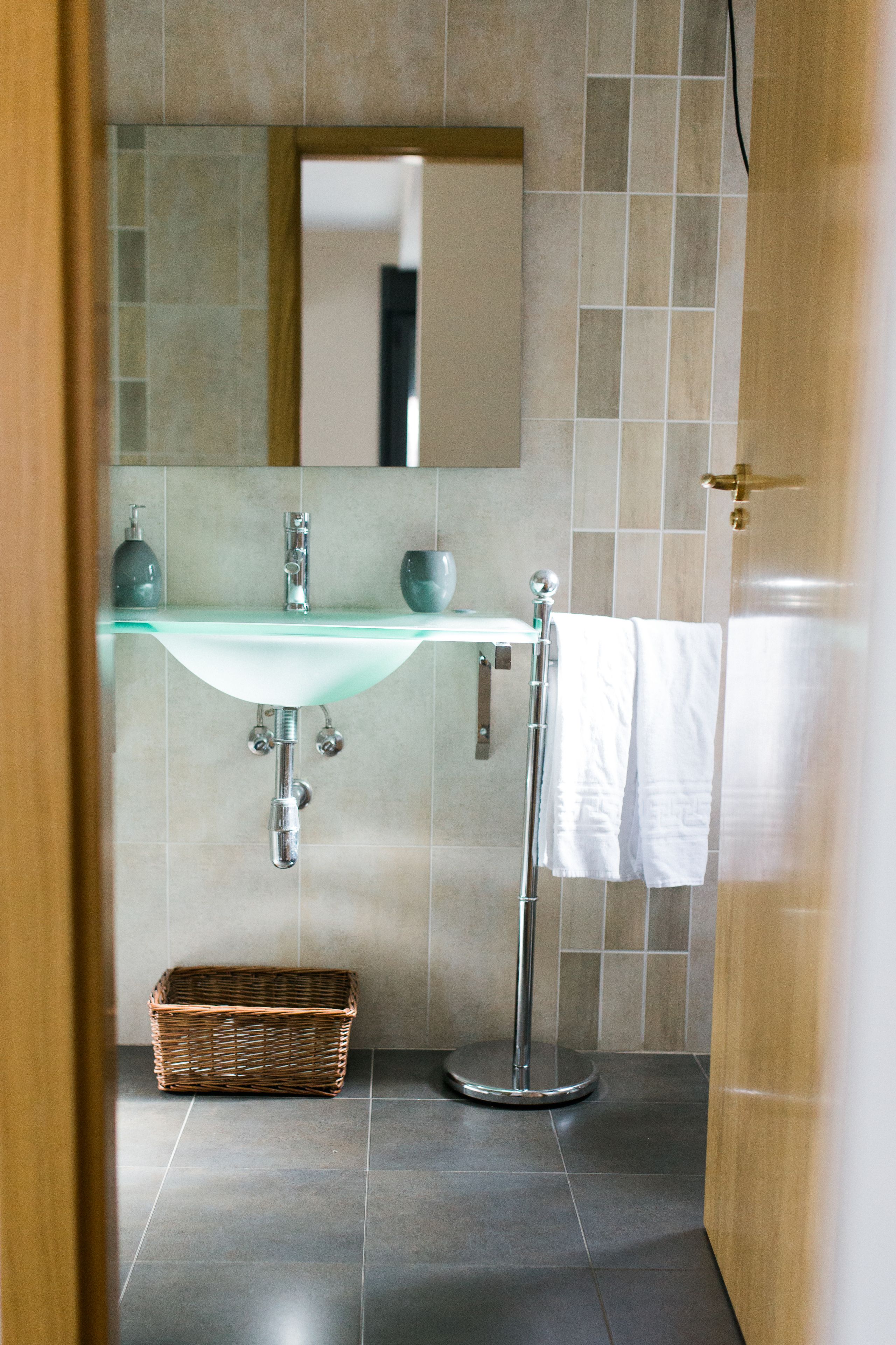 Un baño con lavamanos de vidrio, un espejo, toallero y una cesta de mimbre.