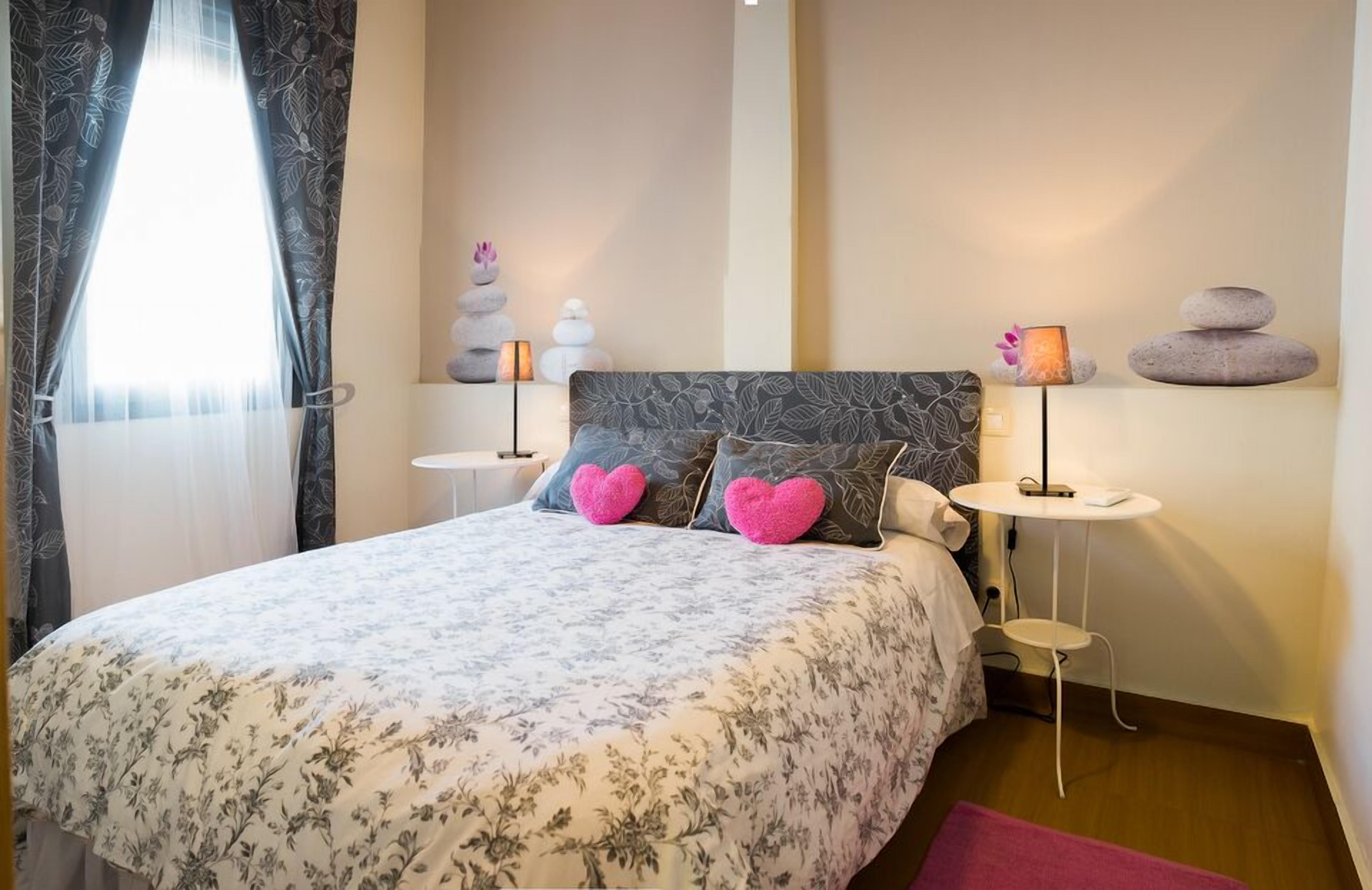 Camera da letto con copriletto floreale, due cuscini a forma di cuore, una testiera con motivo a foglia, due comodini con lampade e una parete con pietre a secco e una piccola pianta.