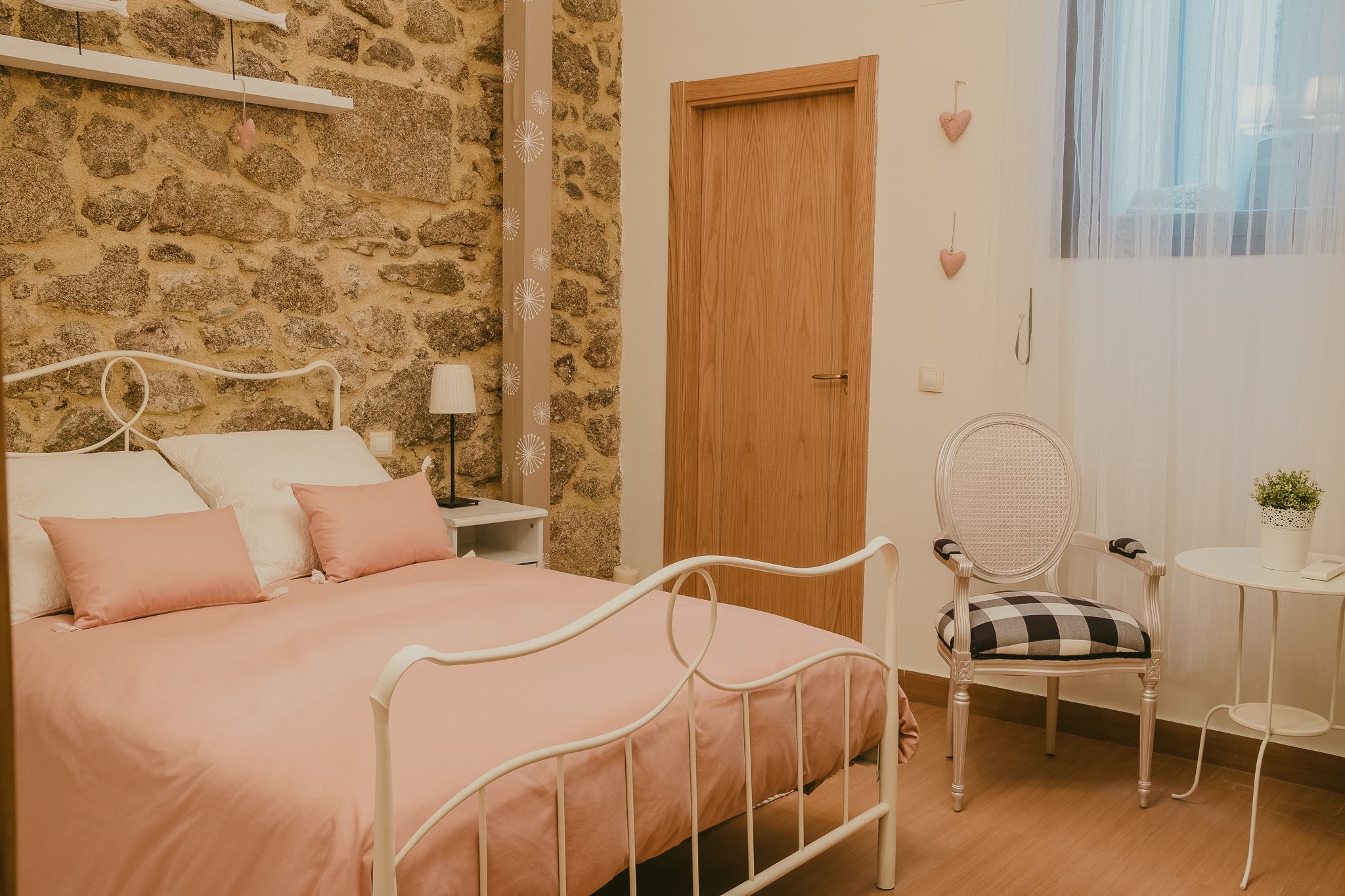 A bedroom with a stone wall, a wooden door, a bed with a metal frame, a chair, a small table with a plant, and a bedside lamp.