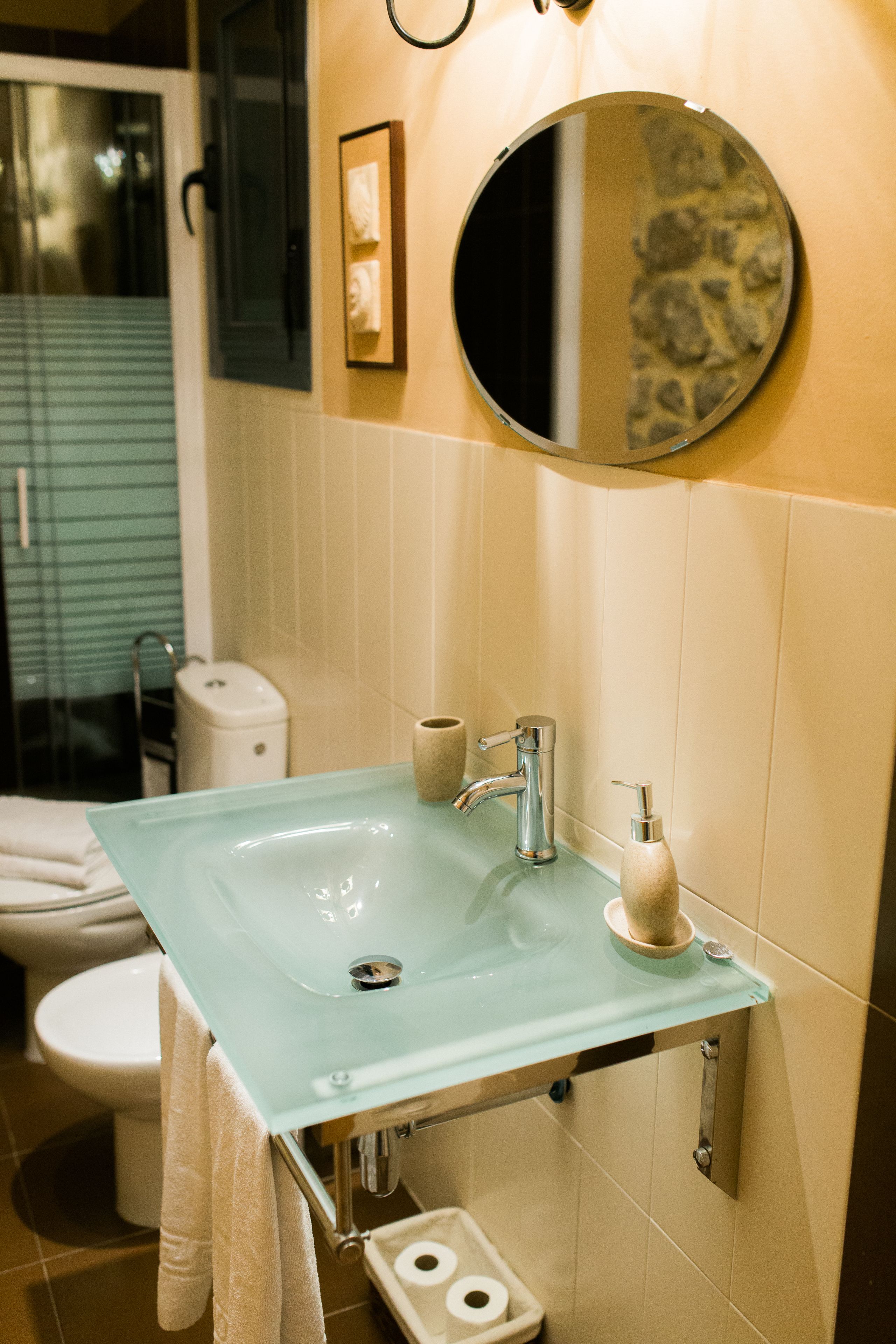 Un bagno con un lavabo in vetro, uno specchio rotondo, un water e un porta asciugamani.