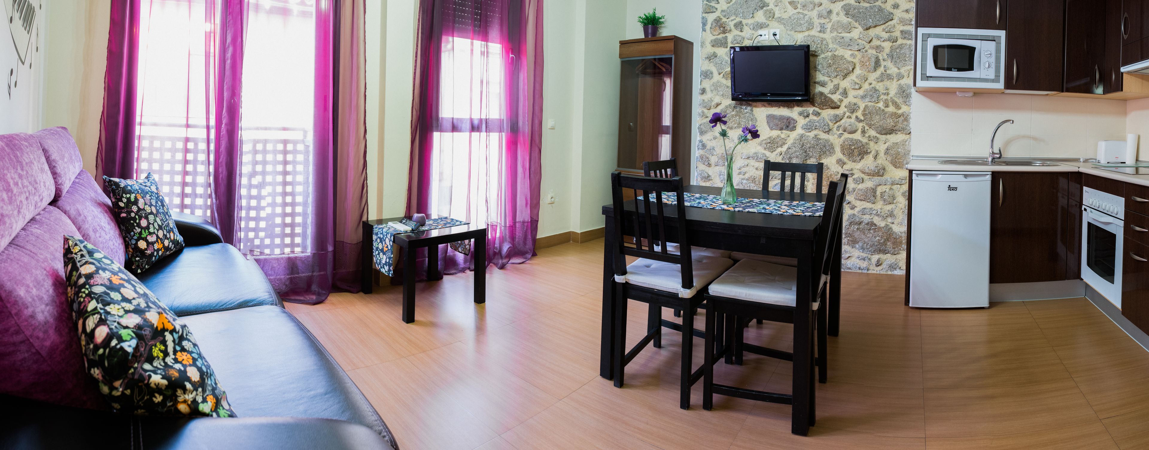 A compact living space with a sofa, dining table, chairs, a small kitchen area with a microwave, and a stone wall.