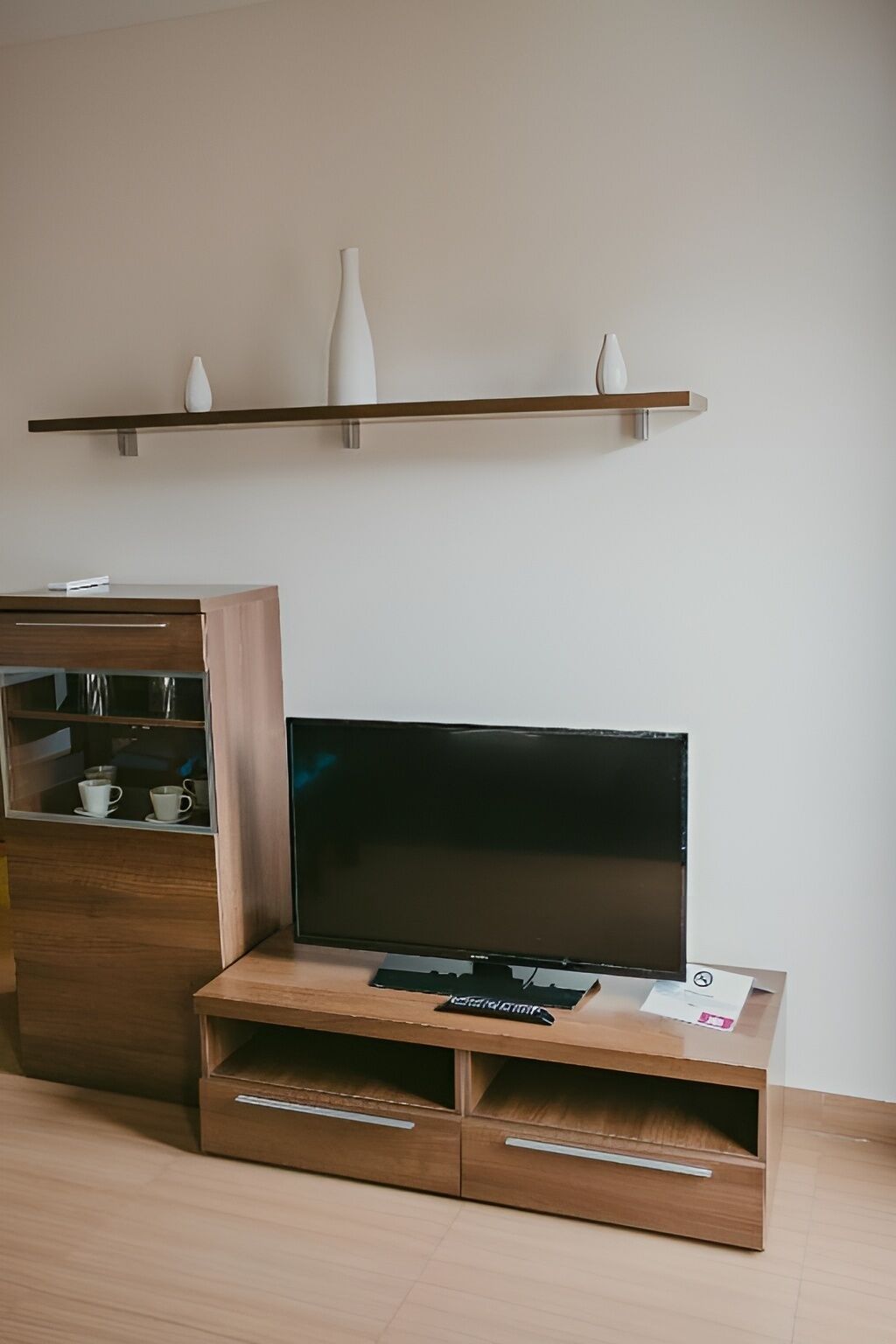 Una sala moderna con un televisor de pantalla plana sobre un soporte de madera, un mueble pequeño con tazas y una estantería con jarrones.