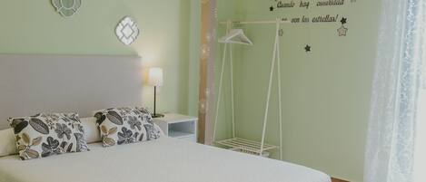 Una cama bien hecha con almohadas estampadas, una mesita de noche con lámpara, y una pared decorada con estrellas y lunas adhesivas.