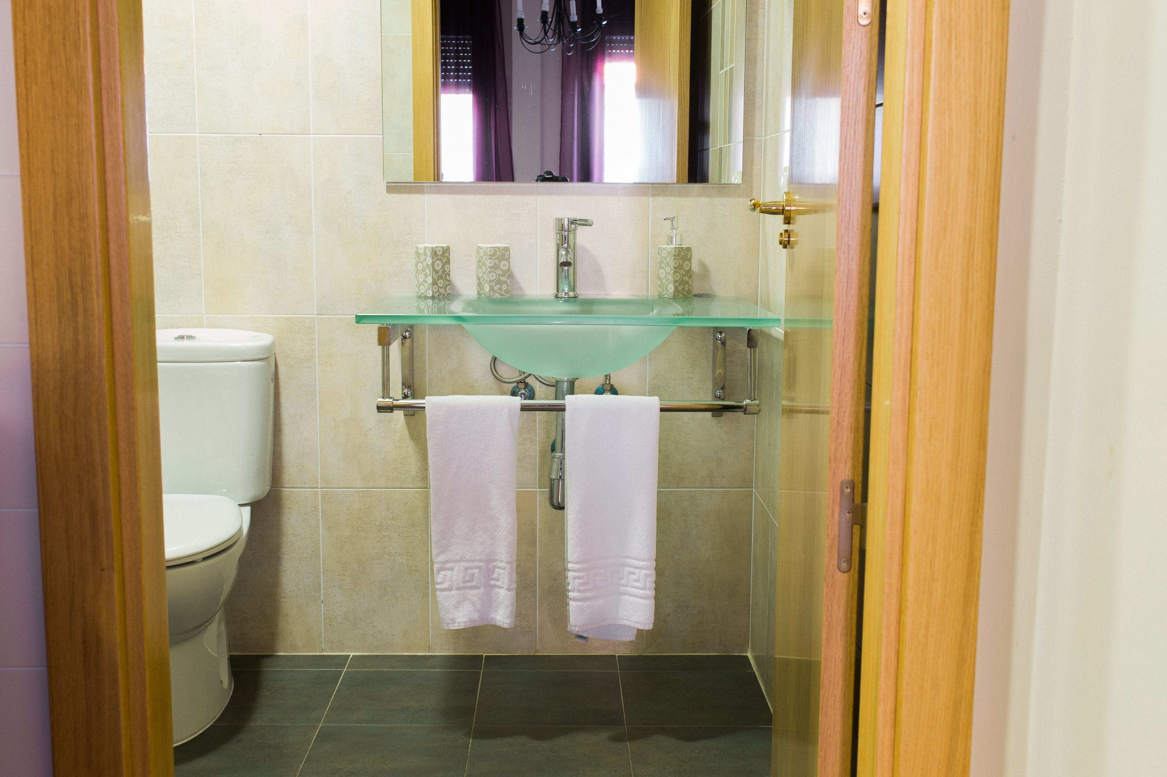Un baño con un lavabo de vidrio, dos toallas blancas, un inodoro y un espejo sobre el lavabo.