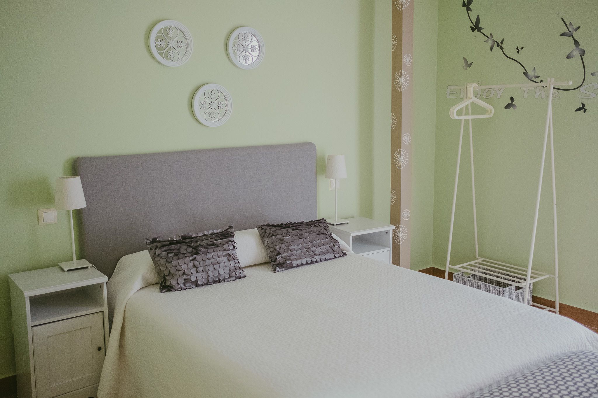 Una cama bien hecha con cabecera gris, dos mesitas de noche con lámparas y placas decorativas en la pared.