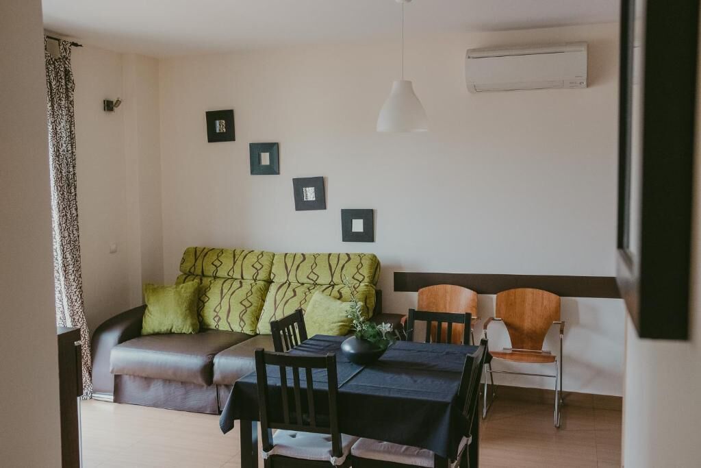 A compact living space with a sofa, a dining table, and chairs. There are framed pictures on the wall, a hanging light fixture, and an air conditioning unit.