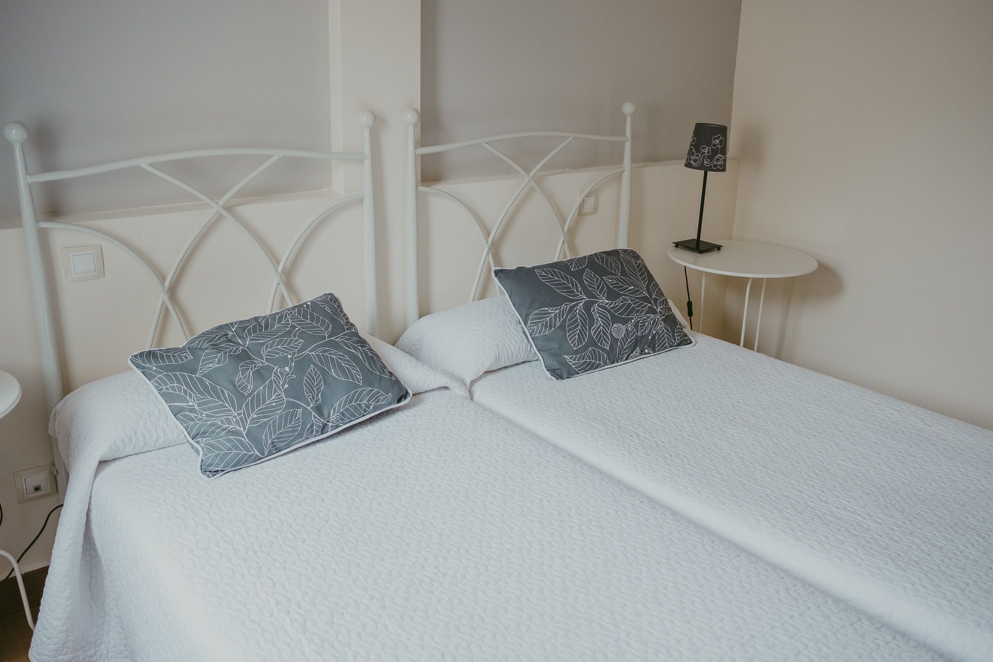 A double bed with white bedding and leaf-patterned pillows, a white metal headboard, and a small bedside table with a lamp.