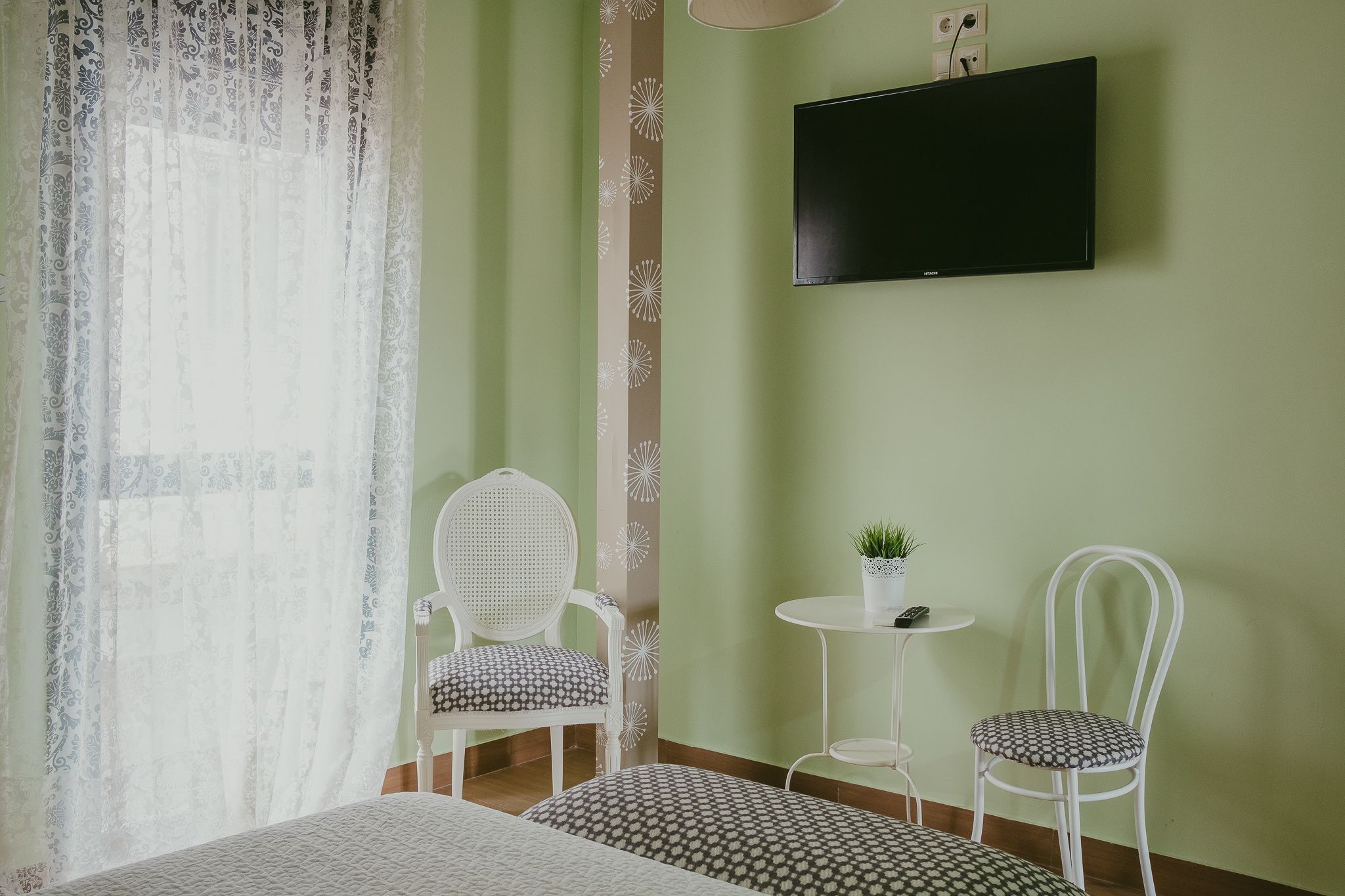 A room with a bed, two chairs, a small table with a plant, and a TV mounted on the wall.