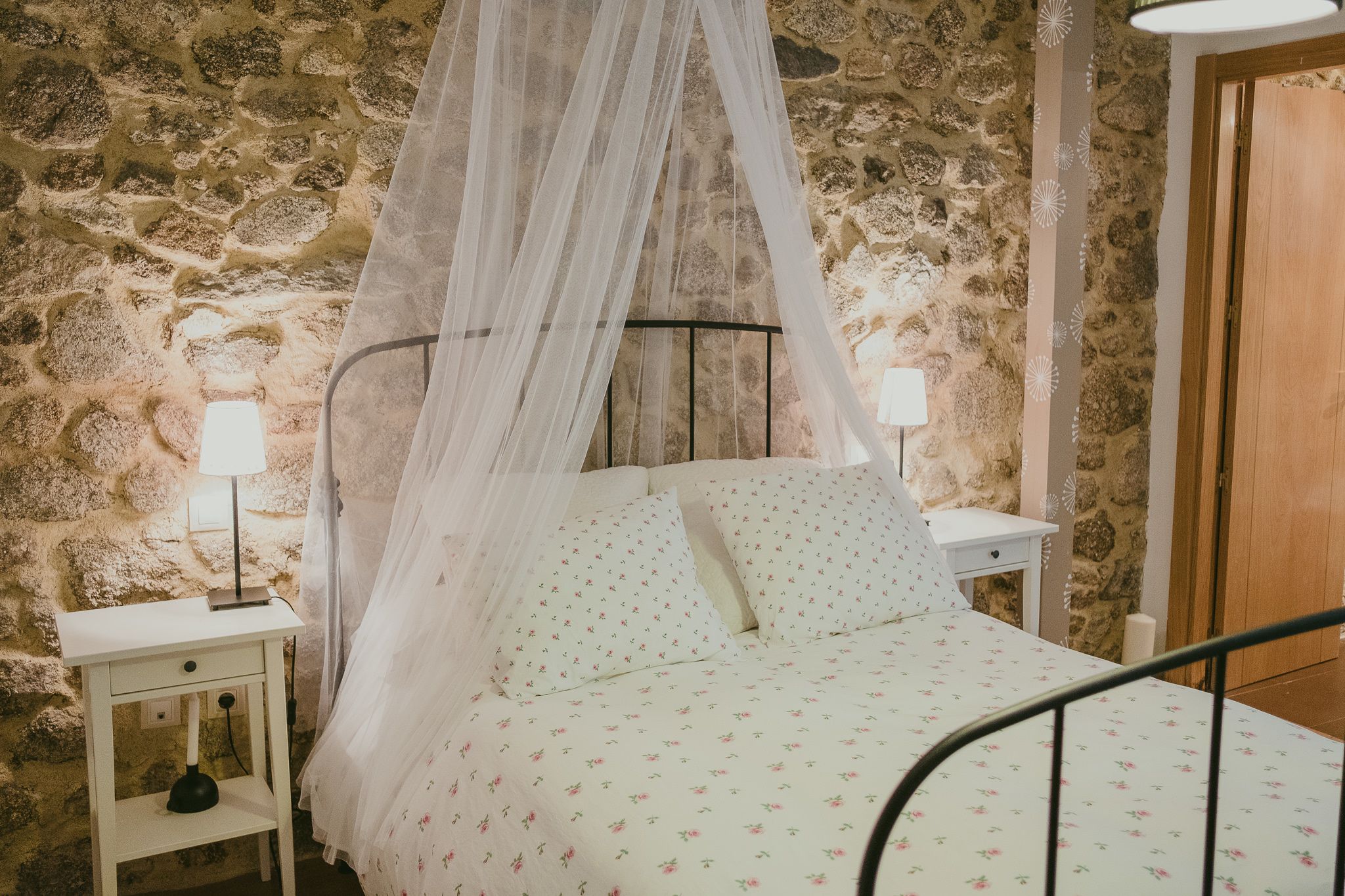 Un dormitorio con pared de piedra, cama con dosel, mesita de noche con lámpara y puerta de madera.