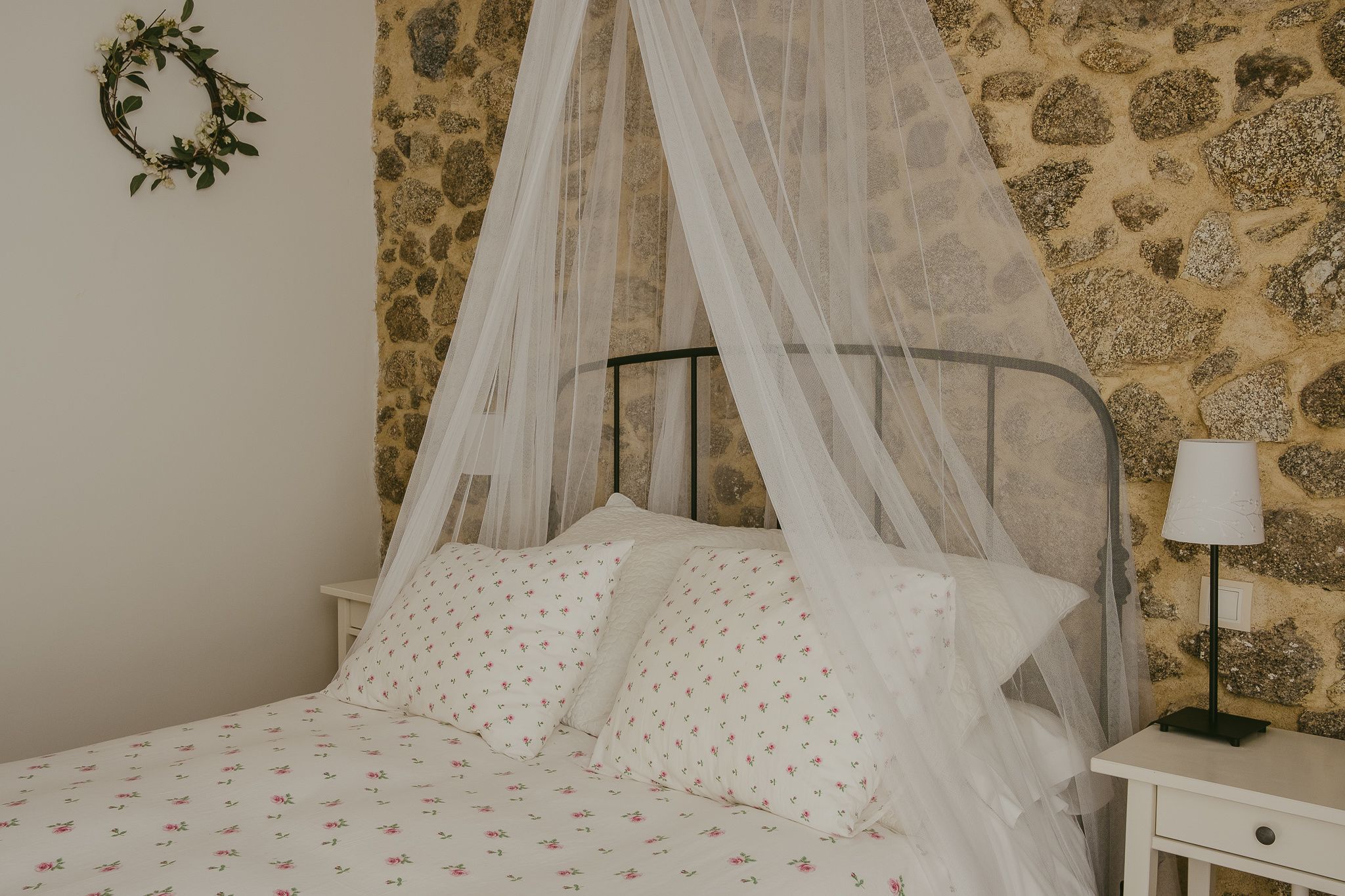 Una cama con dosel, ropa de cama blanca con motivos florales, una mesita de noche con lámpara y una pared de piedra.