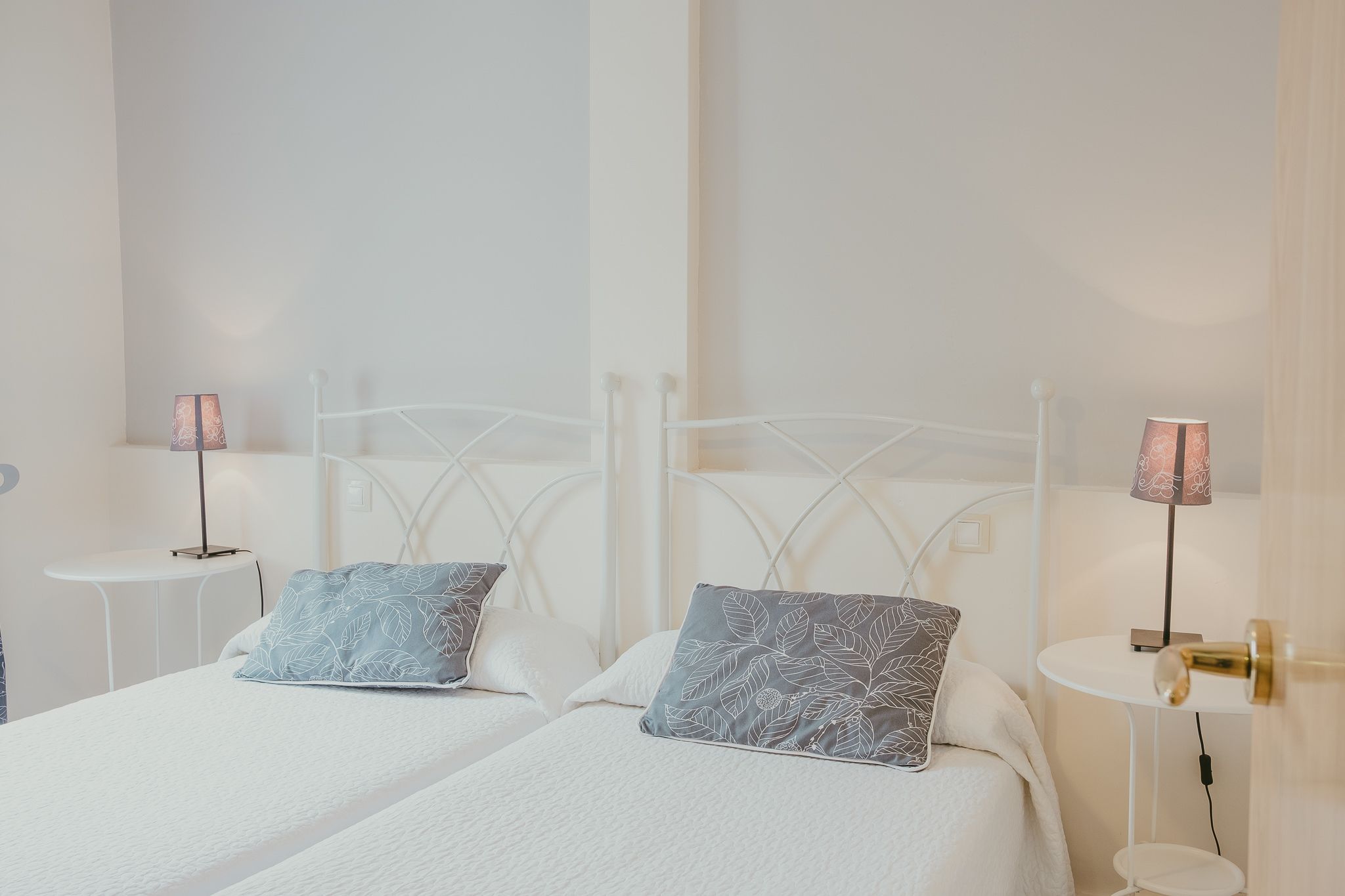 Un dormitorio limpio y moderno con dos camas, cada una con cabecero blanco y almohadas a juego. Hay mesitas de noche con lámparas y una pequeña mesa entre las camas.