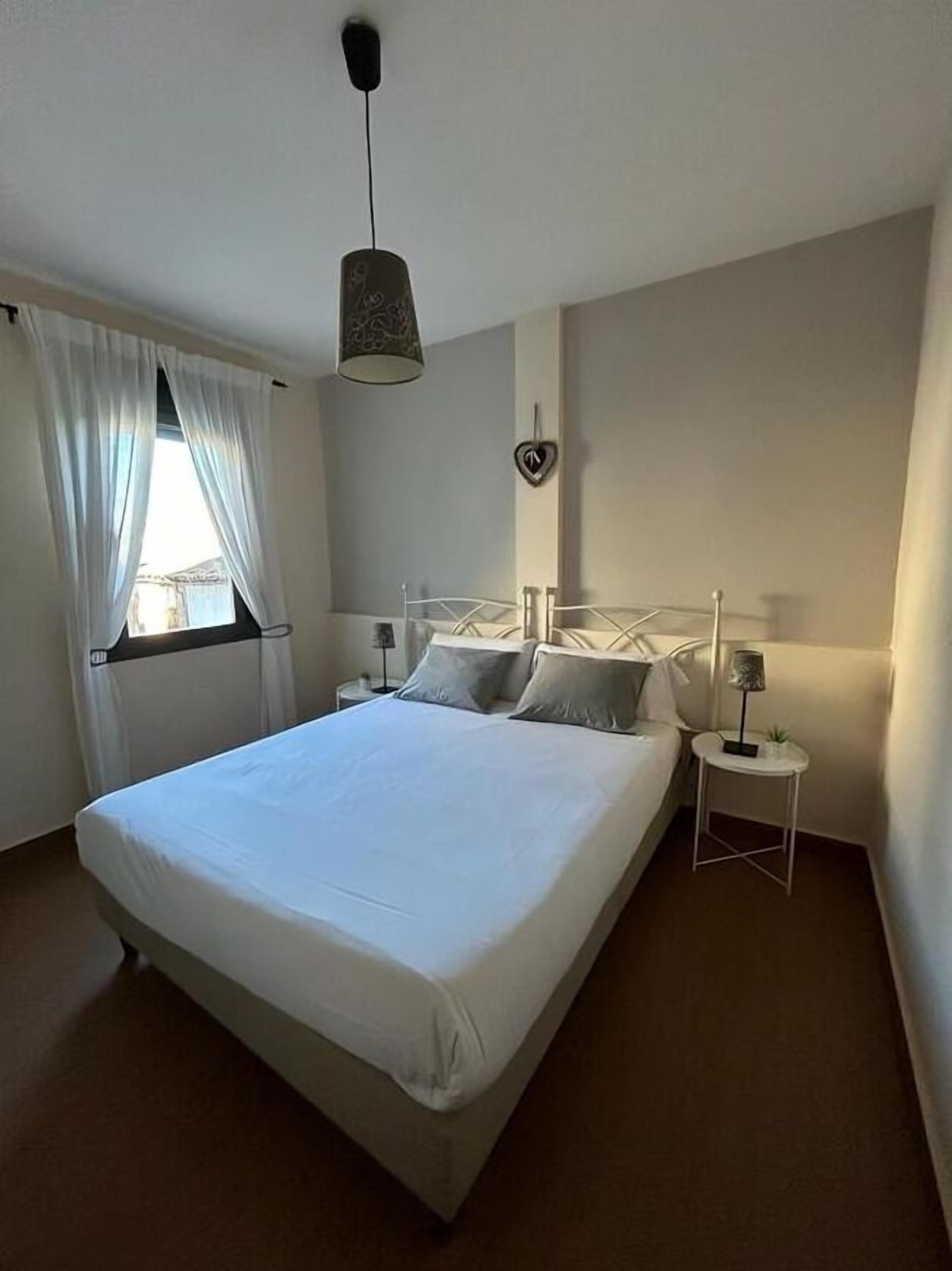 A neatly made bed in a room with a single bedside table, a hanging lamp, and a window with curtains.
