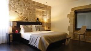 Un dormitorio moderno con pared de piedra, una cama con colcha color beige, dos lámparas de noche, una mesita de noche y una silla.