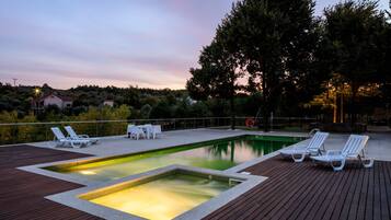 A rectangular swimming pool with a wooden deck, surrounded by white lounge chairs and a table.