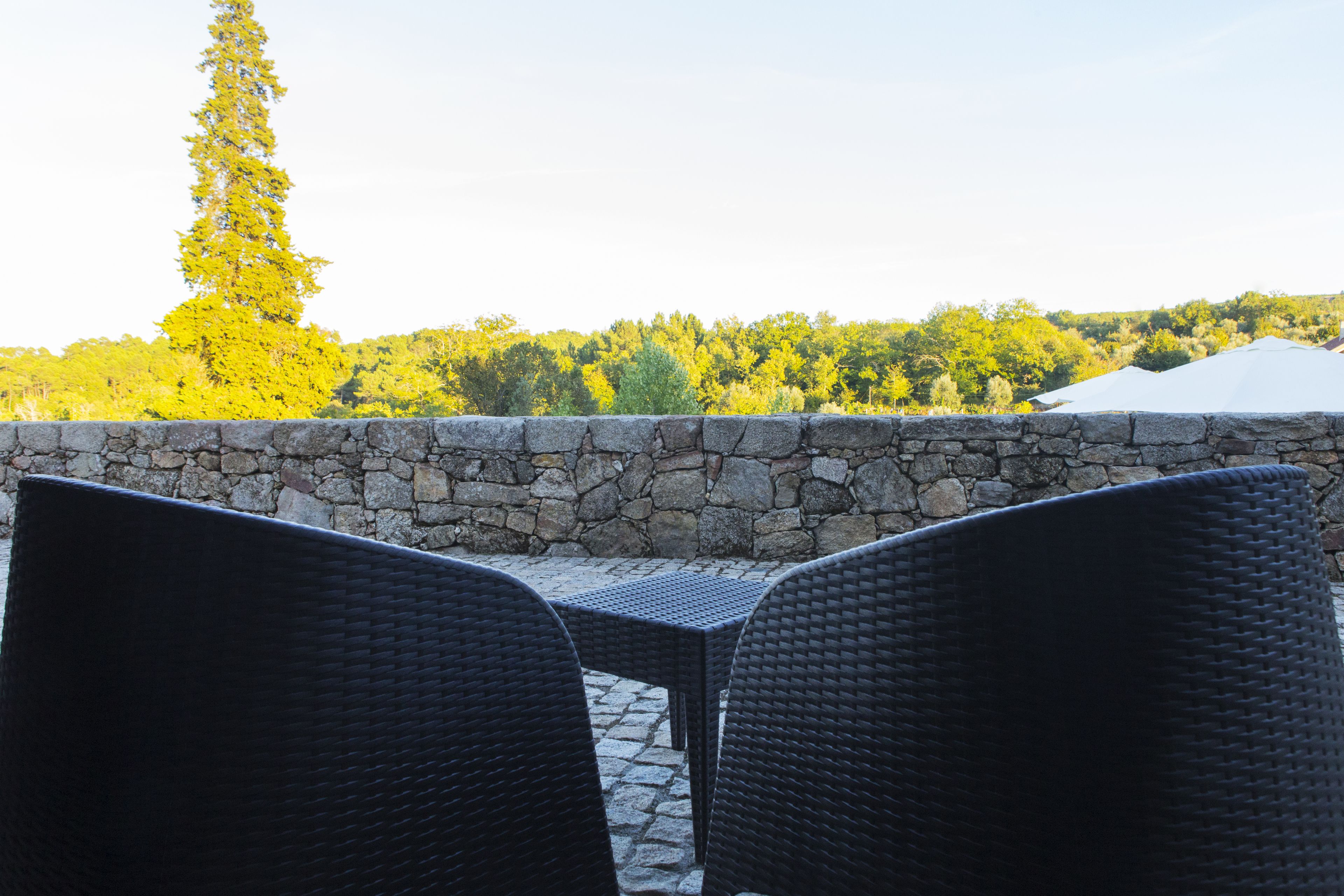 A stone wall with wicker chairs and a table, overlooking a forested hillside.