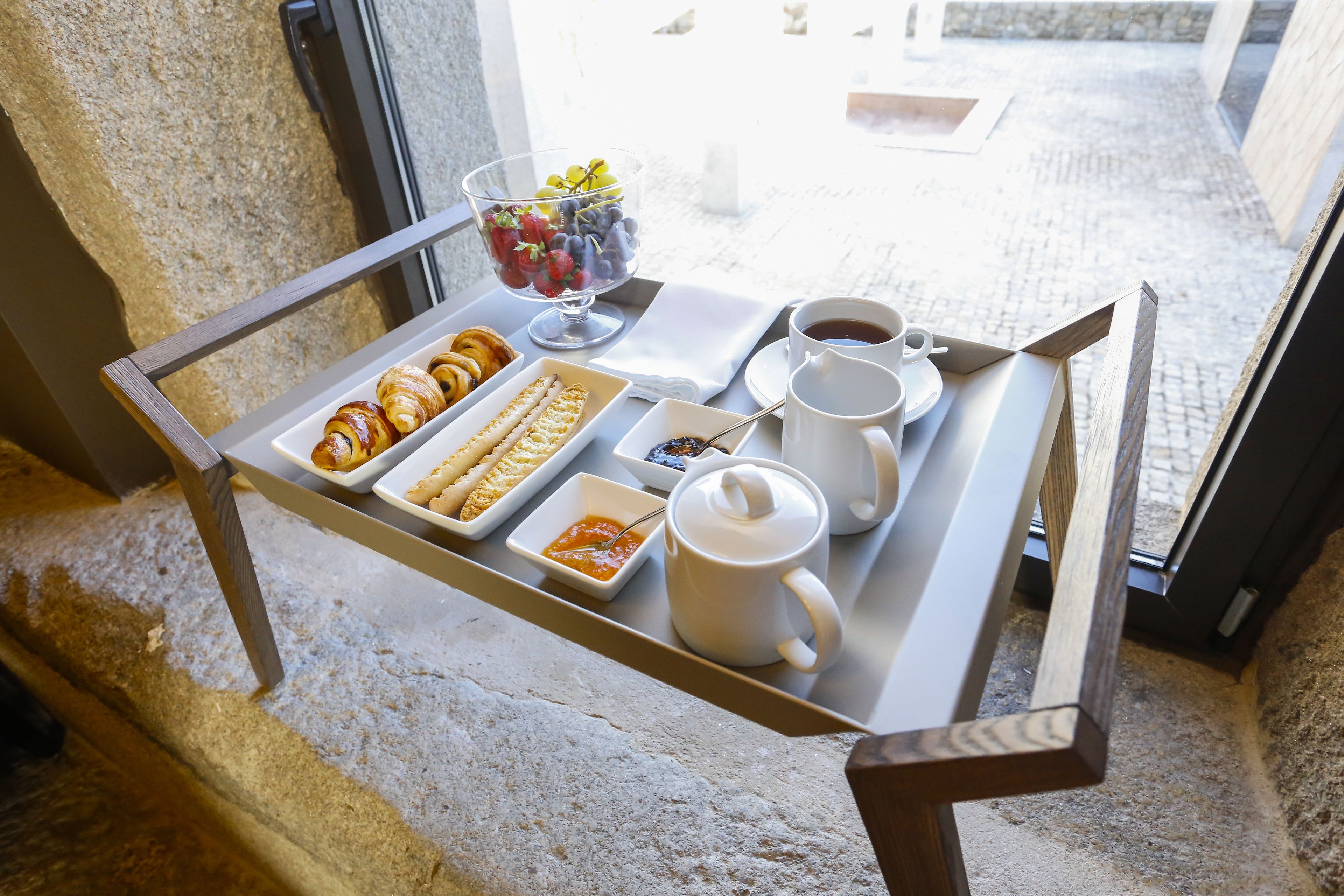 Una bandeja con pasteles, un frutero, una taza de café y una tetera sobre una mesa junto a una ventana.