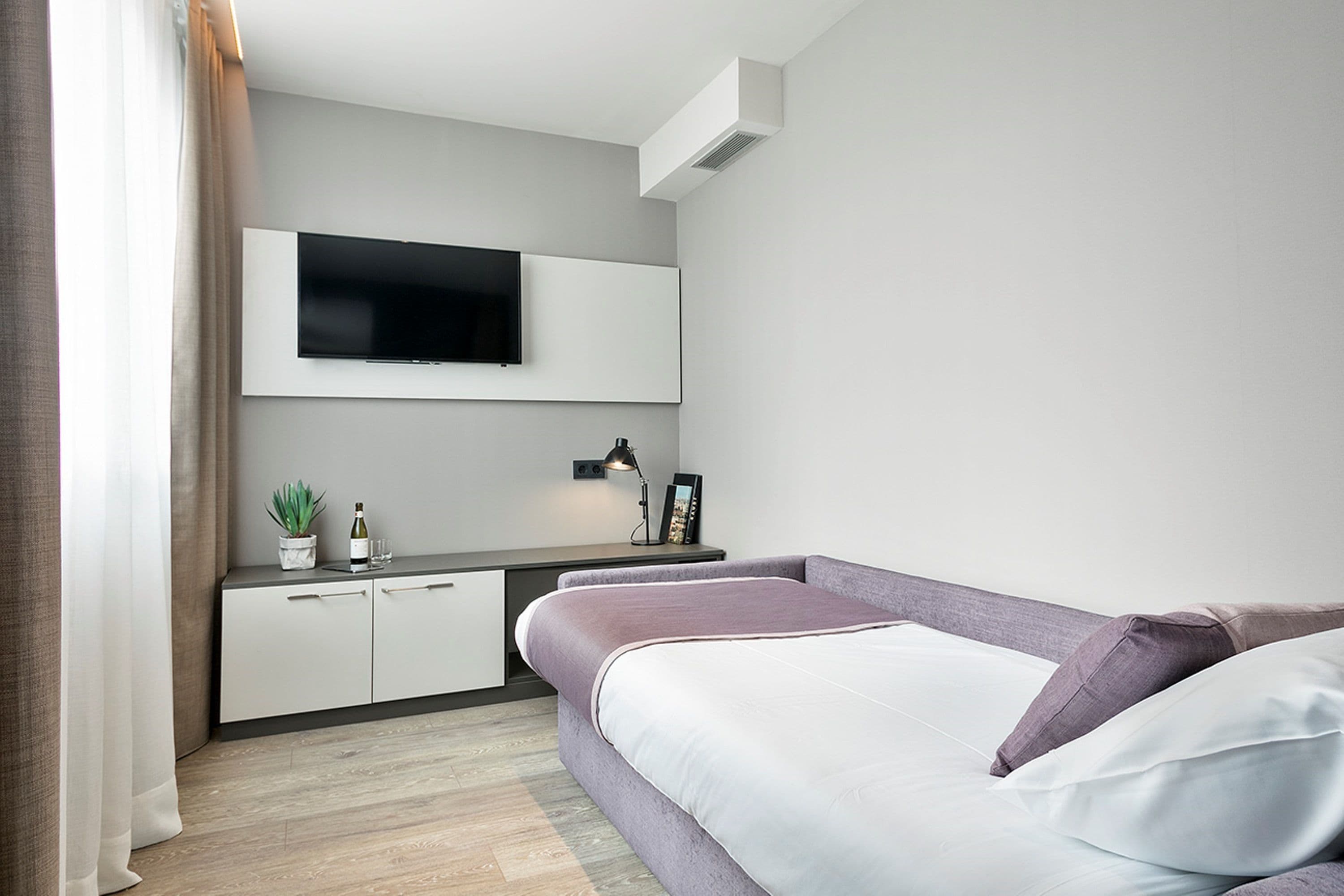 A modern bedroom with a bed, a TV mounted on the wall, a bedside table with a lamp, and a small plant.