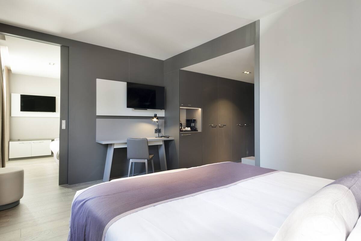 A modern hotel room with a large bed, a desk with a chair, a flat-screen TV mounted on the wall, and a small kitchenette area.