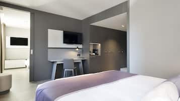 A modern hotel room with a large bed, a desk with a chair, a flat-screen TV mounted on the wall, and a small kitchenette area.