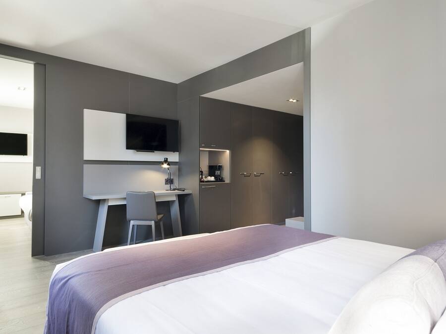 A modern hotel room with a large bed, a desk with a chair, a flat-screen TV mounted on the wall, and a small kitchenette area.