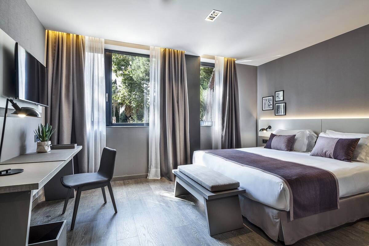 A modern hotel room with a bed, desk, chair, and large windows with curtains.