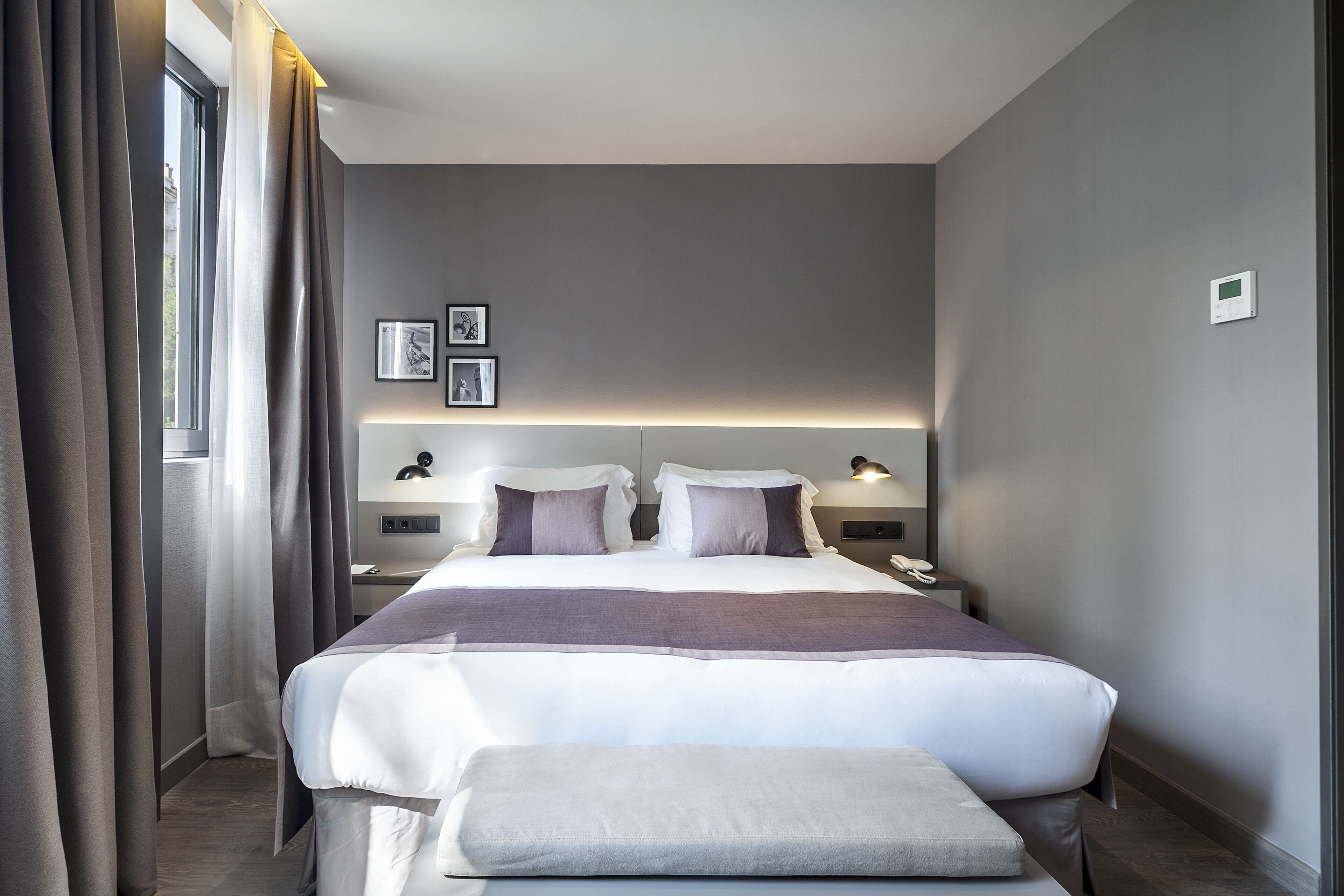 A modern hotel room with a large bed, bedside tables, a nightstand, and a window with curtains.