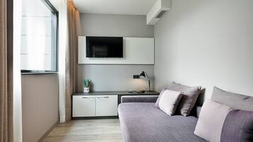 Ett modernt sovrum med en grå soffa, en platt-TV som är monterad ovanför ett nattduksbord och ett fönster med gardiner.