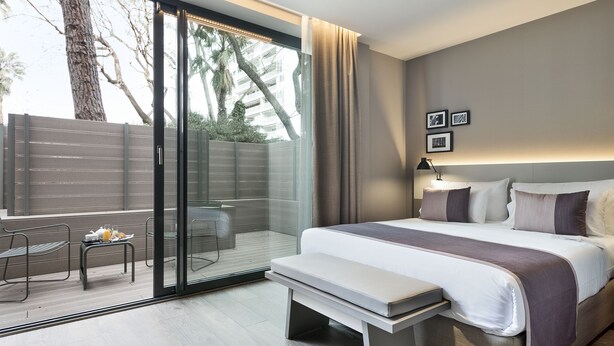 A modern hotel room with a large bed, a bench, a small table with a tray of breakfast, a sliding glass door to a balcony, and a city view. - Hotel Upper Diagonal (Barcelona)