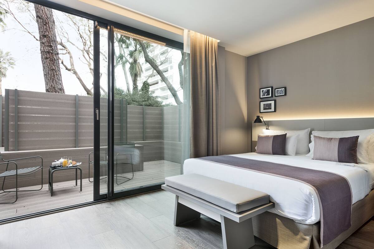 A modern hotel room with a large bed, a bench, a small table with a tray of breakfast, a sliding glass door to a balcony, and a city view.