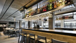 A modern bar area with a long counter, multiple seating options, and a variety of liquor bottles displayed on shelves. - Hotel Upper Diagonal (Barcelona)