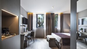 A modern hotel room with a bed, desk, and chair, a small table with a plant, and a window with curtains.