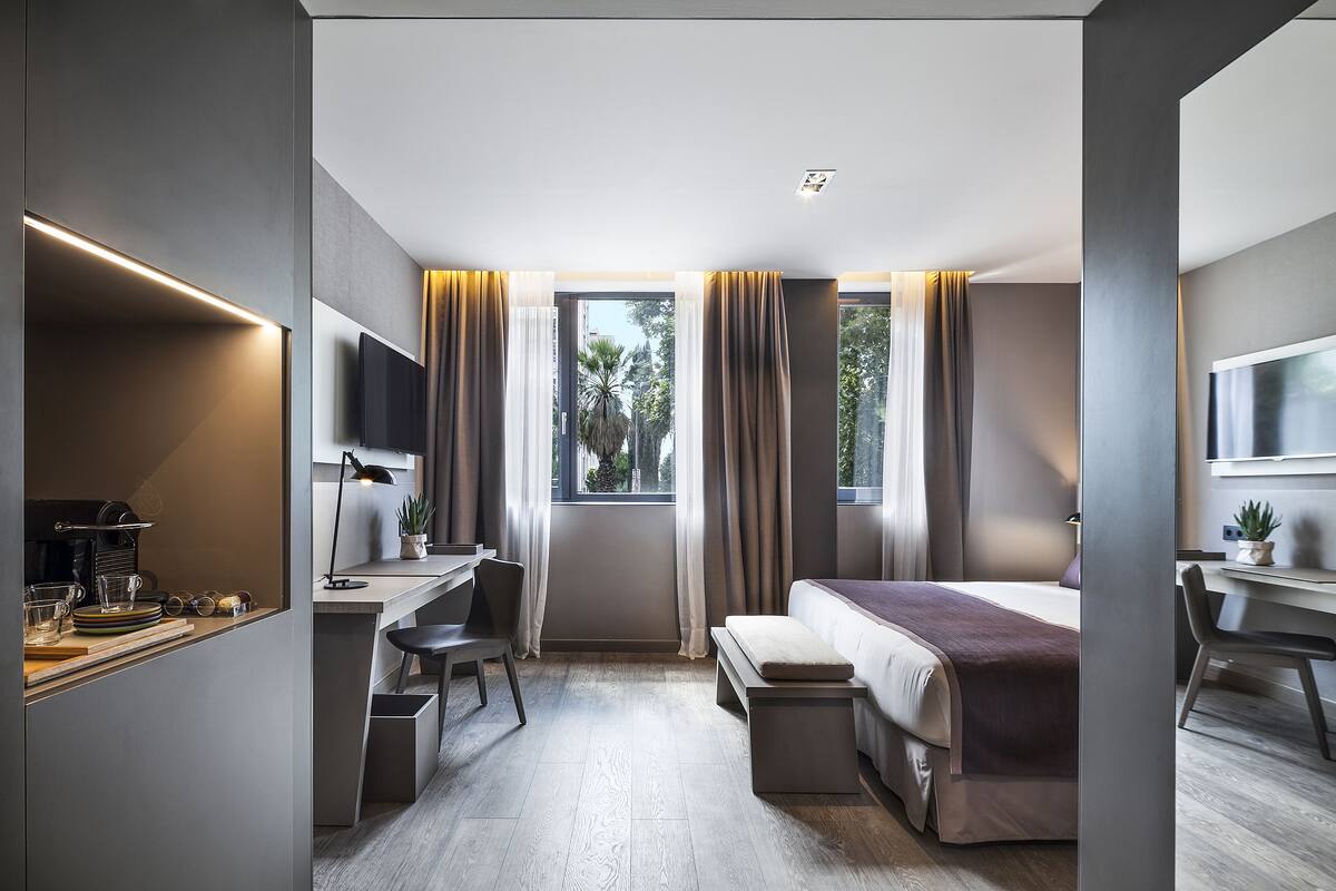 A modern hotel room with a bed, desk, and chair, a small table with a plant, and a window with curtains.