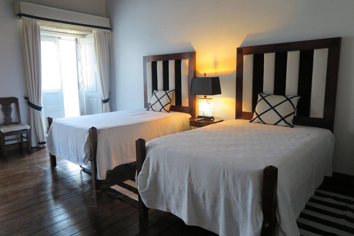 A hotel room with two single beds, a nightstand with a lamp, a wooden chair, and a window with curtains.
