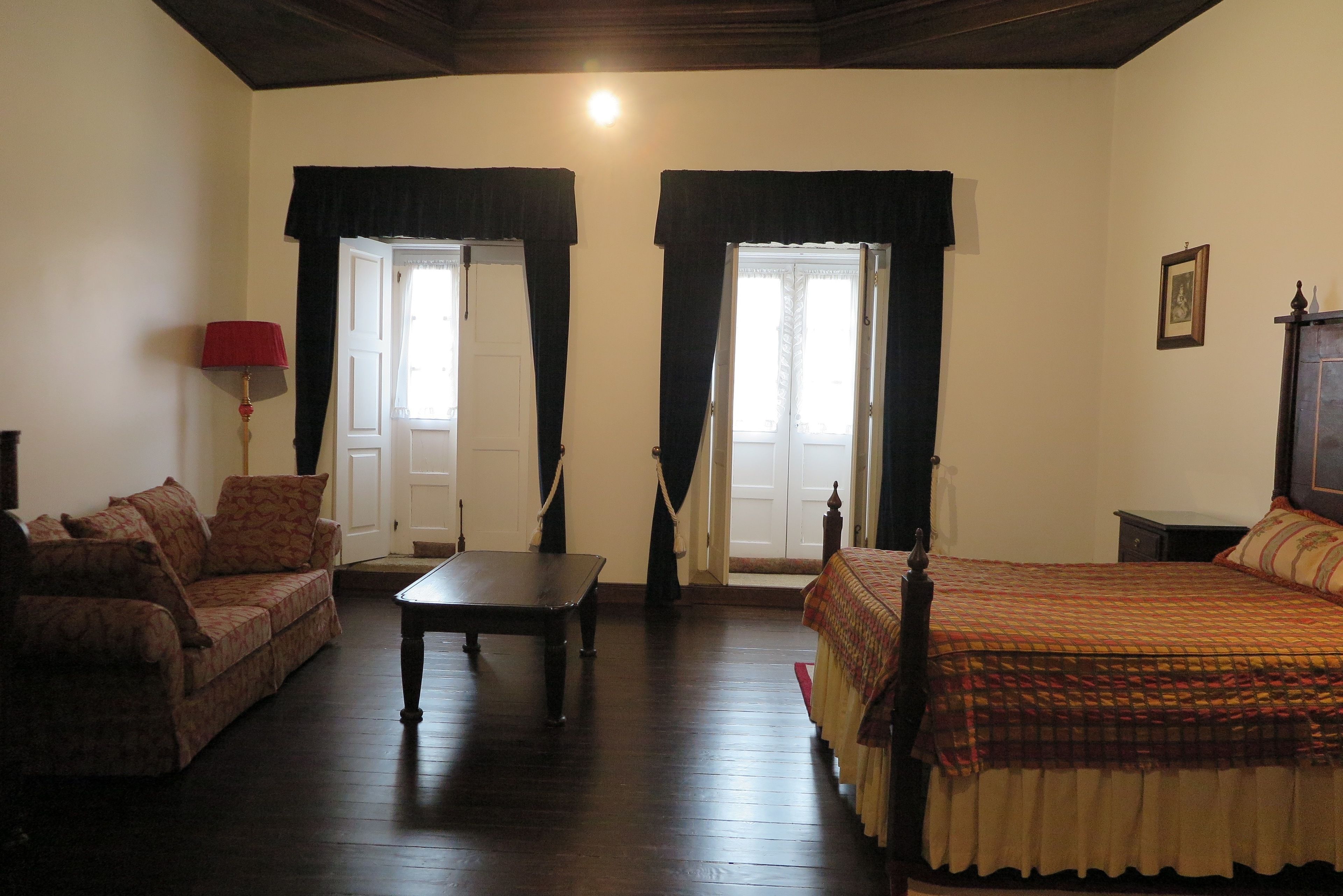 A room with a sofa, a table, a bed, and two windows with curtains.