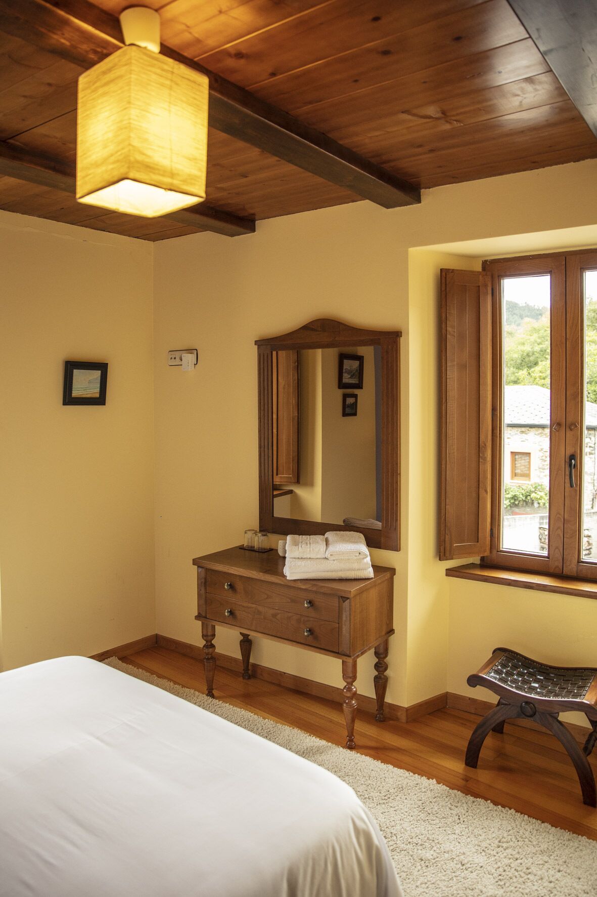 A bedroom with a wooden ceiling, a bedside table with a mirror, a bed, a window with wooden shutters, and a small bench.
