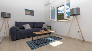 A modern living room with a dark blue sofa, a wooden coffee table, and a blue patterned rug. There is a window with a view of greenery and a clear sky. - Villa Carica (Omis)