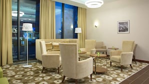 Un hall de hotel con un sofá, sillones, alfombra estampada, un ventanal con cortinas y un cuadro enmarcado en la pared.