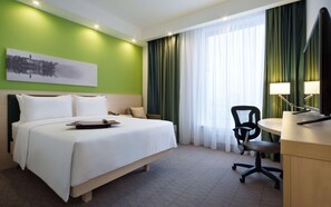 Una habitació d'hotel amb un llit, un escriptori amb un ordinador, una cadira, una làmpada i un quadre a la paret.