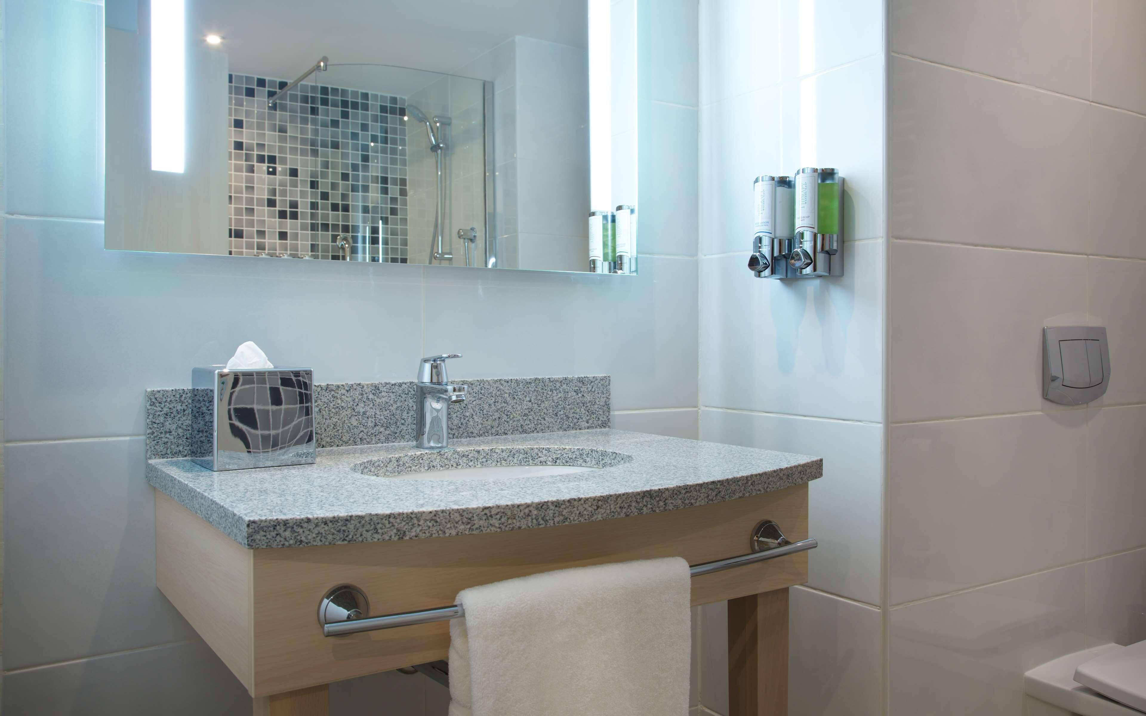 A modern bathroom with a granite countertop sink, a mirror, a towel rack, and a shower area.