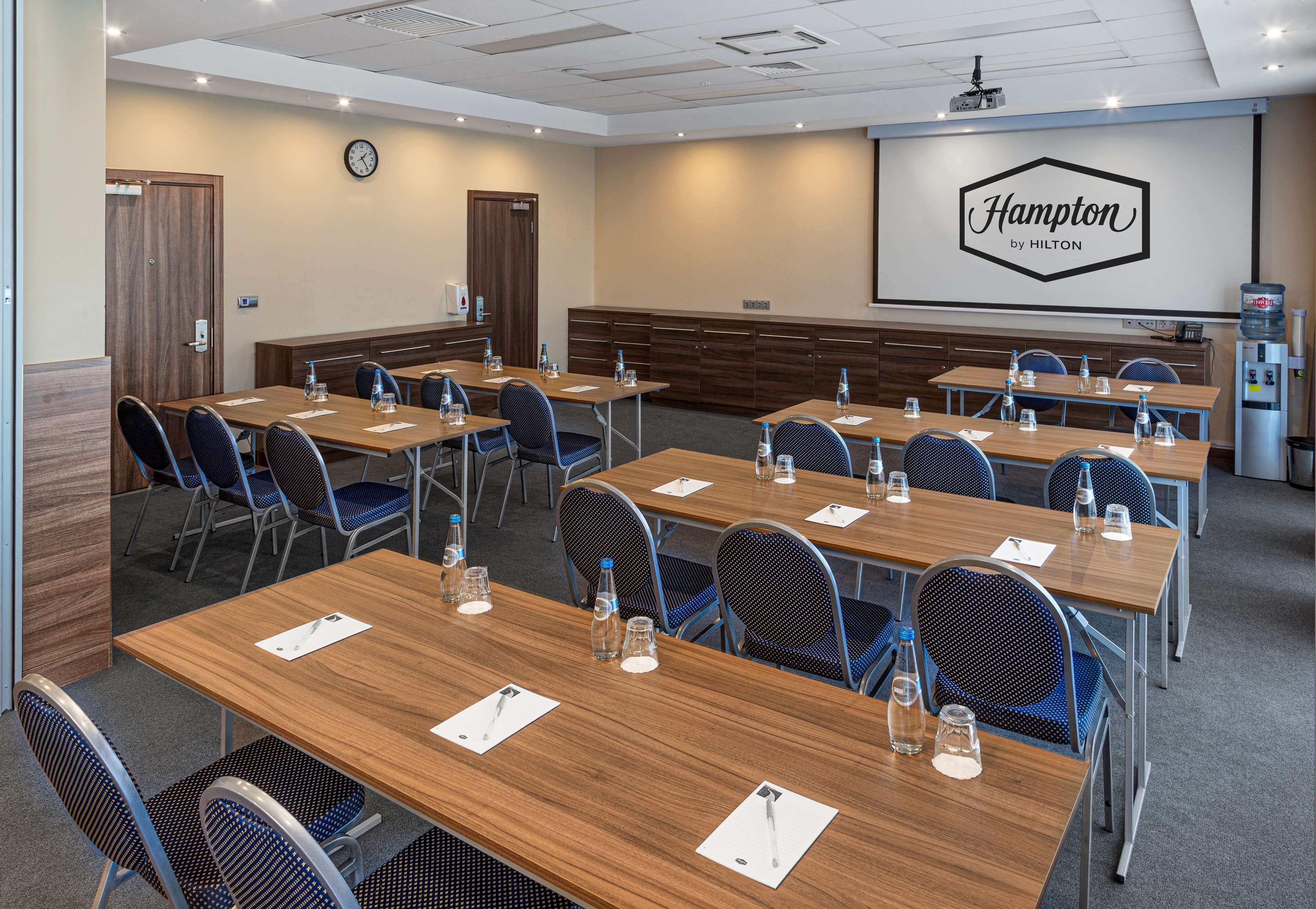 Un salón de conferencias con varias mesas y sillas, un reloj, un dispensador de agua y una pantalla de presentación con el logo de Hampton by Hilton.