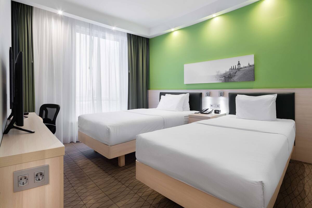 Ein Hotelzimmer mit zwei Betten, einem Schreibtisch, einem Sessel, einem Flachbildfernseher und einem Bild an der Wand.