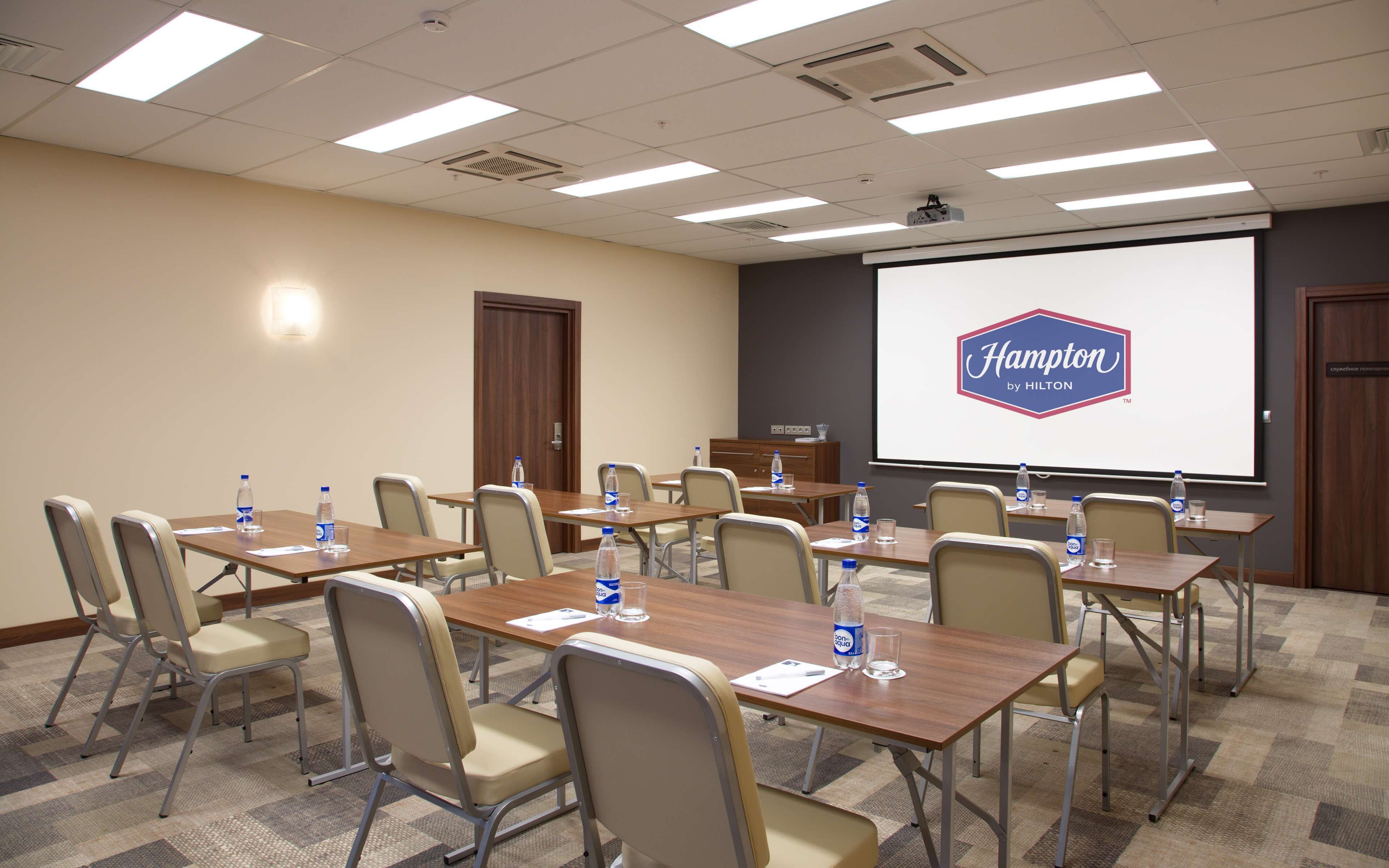 A conference room with tables, chairs, and a projector screen displaying 'Hampton by Hilton'.