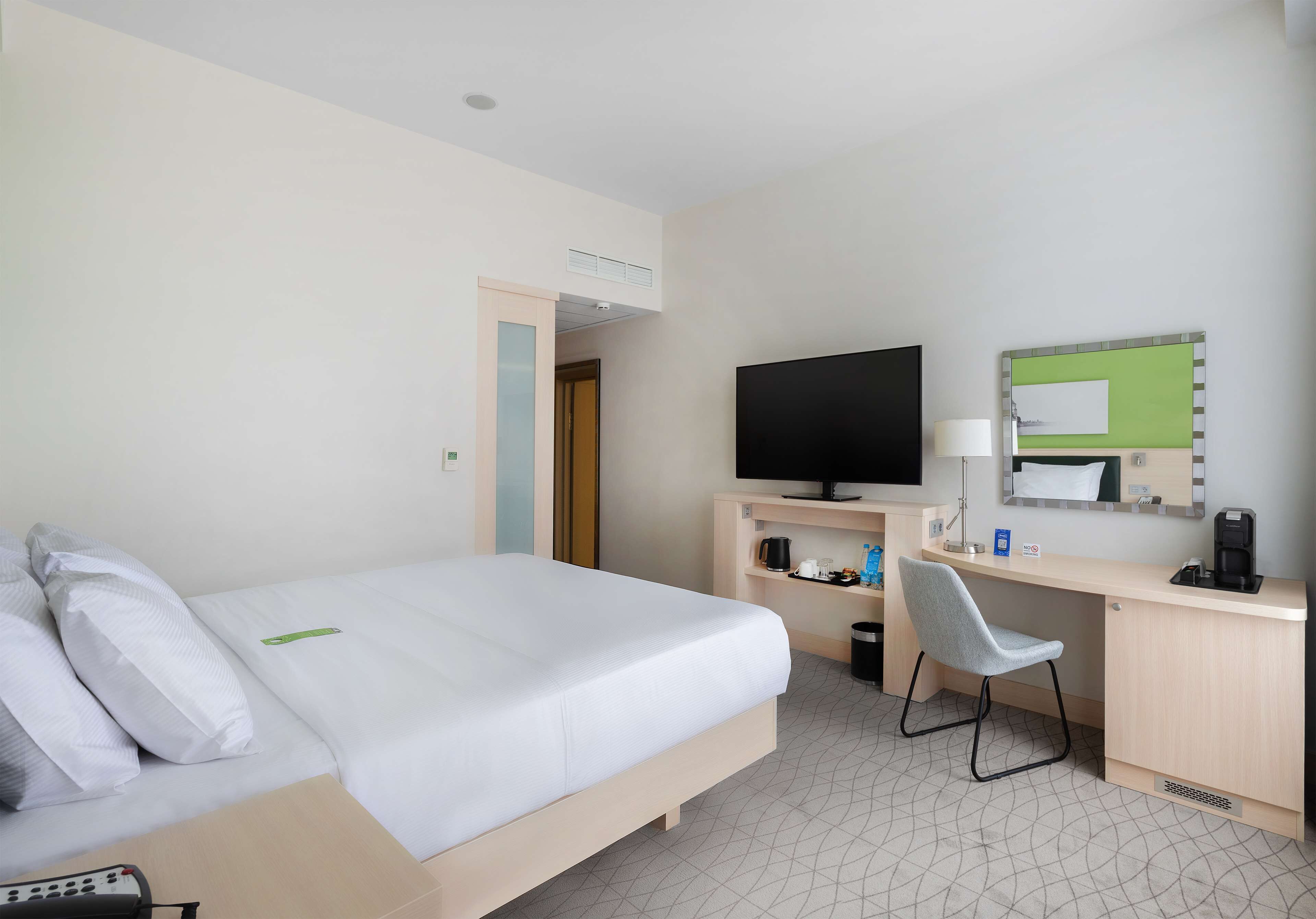A hotel room with a bed, a desk with a computer, a chair, a TV, and a small cabinet with toiletries.