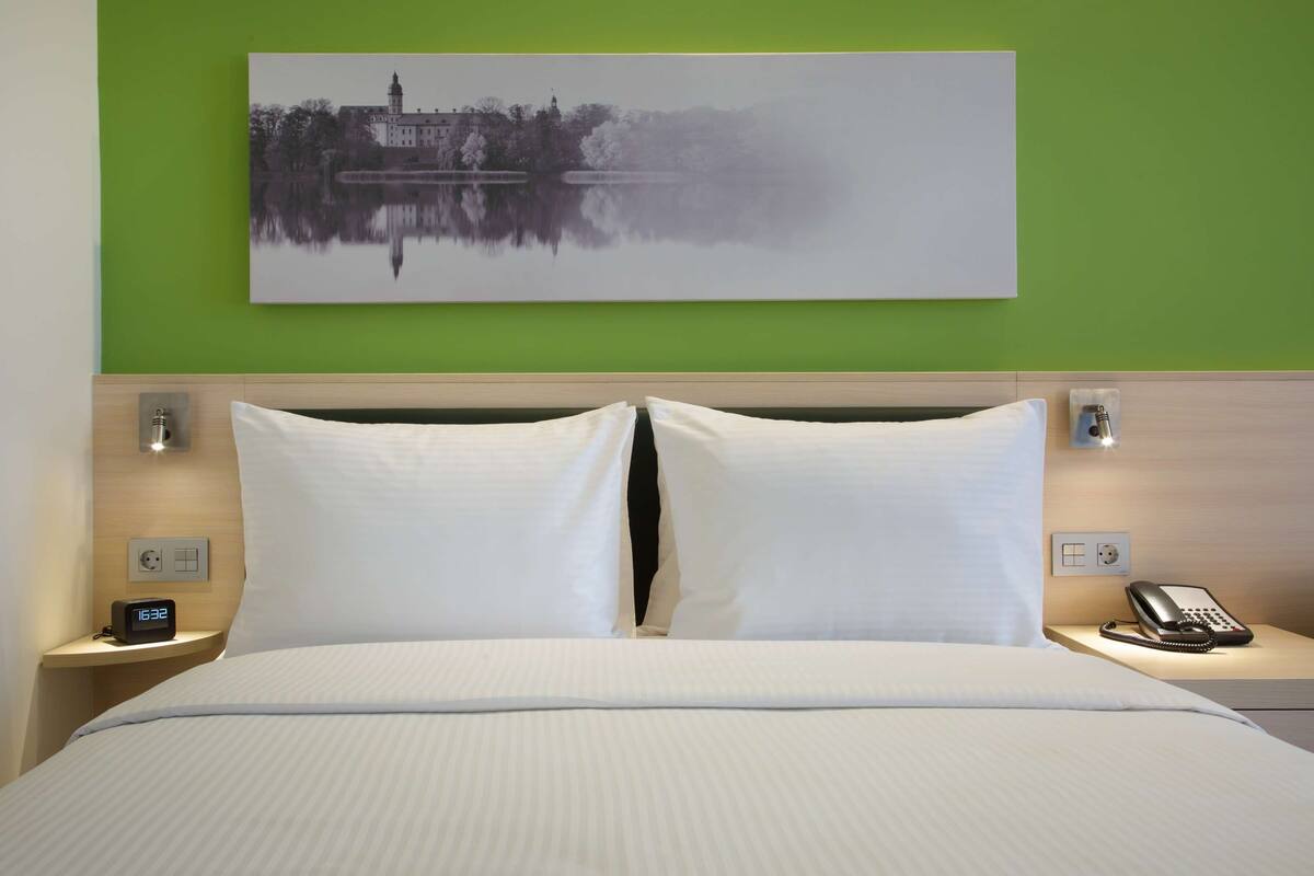 Ein Hotelzimmer mit einem Bett, zwei Nachttischen, einem Schreibtisch und einem Landschaftsbild an der Wand.
